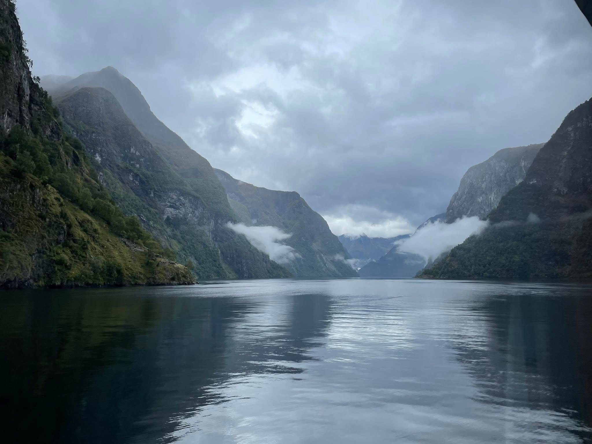 Sognefjord with calm water reflecting steep cliffs and low clouds, view from a boat in Norway.