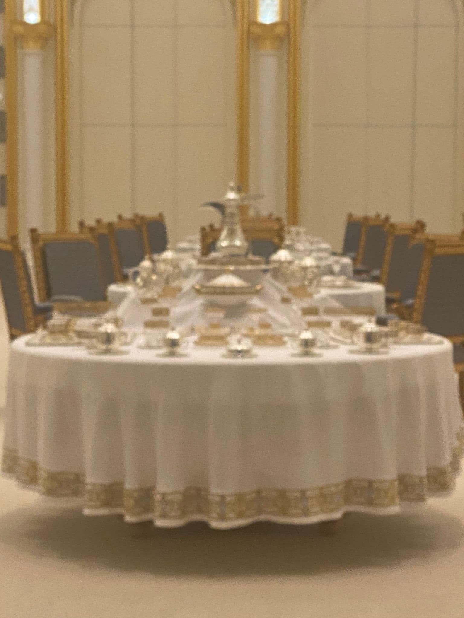 Round formal dining table set with silver service in an ornate palace dining room, gilded chairs and paneling visible.