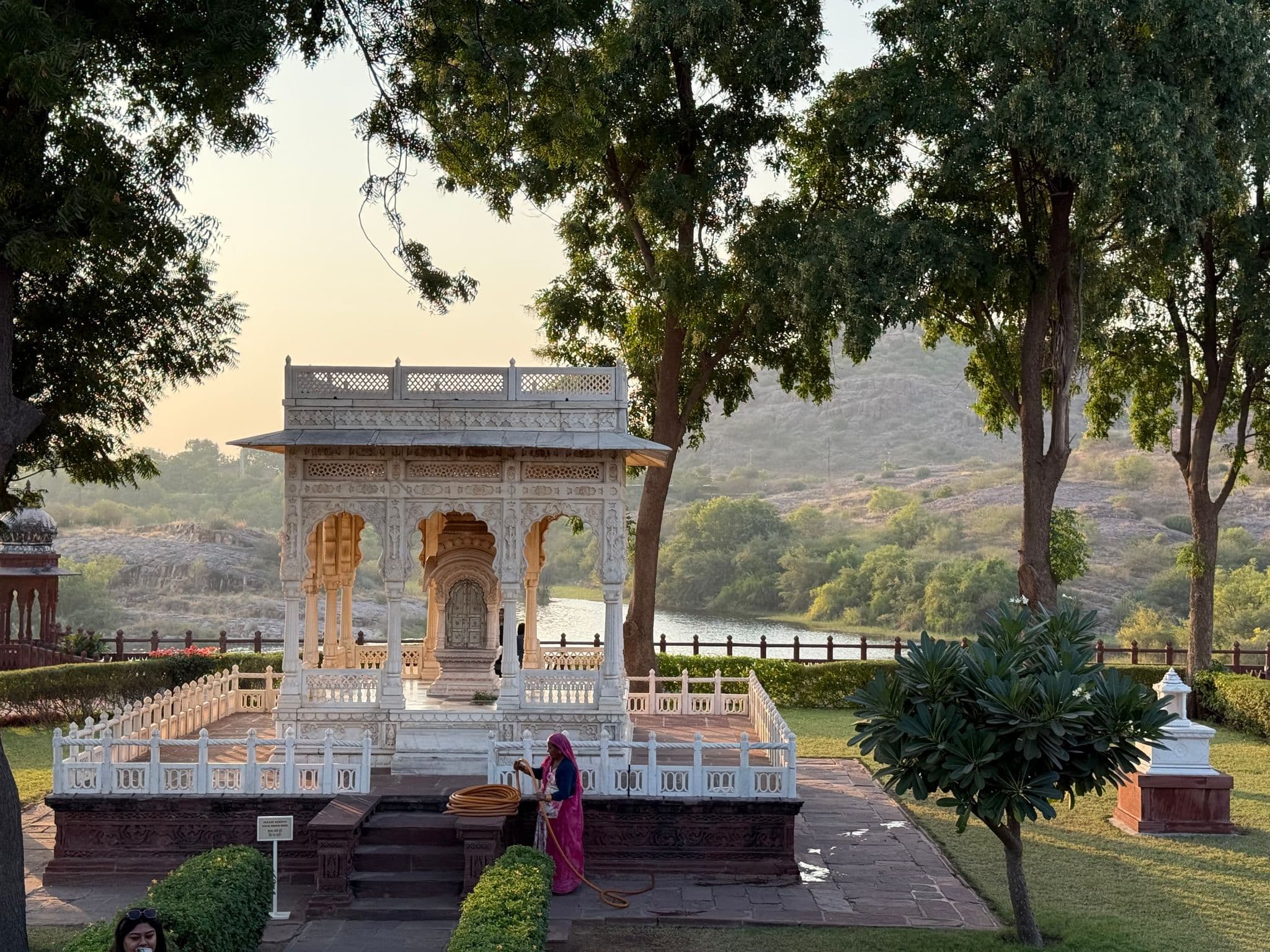 Ornate white marble pavilion by a lakeside with sunlight through trees and a woman in bright clothing tending the garden, Rajasthan, India.