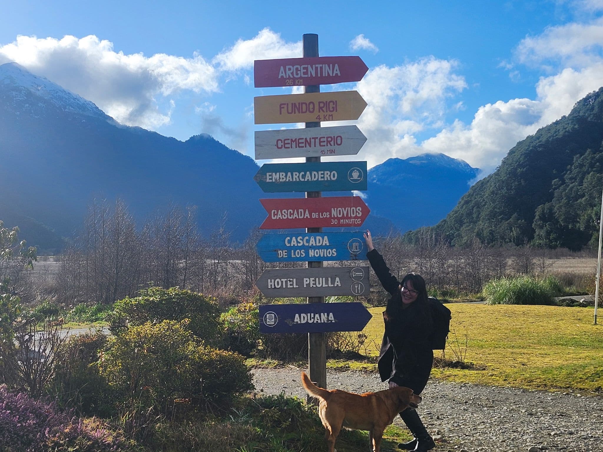 Colorful wooden signpost naming Peulla and Cascada de los Novios, with a person and a dog beside it and mountains in Chile.