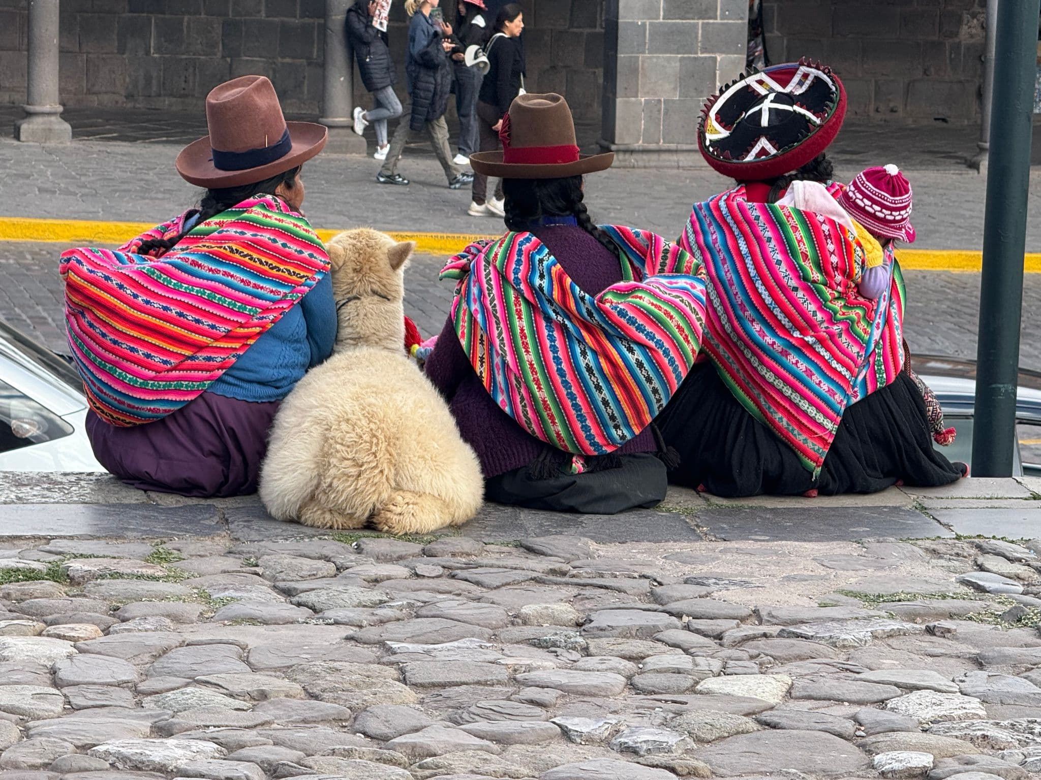 Three women in colorful Andean shawls sitting on stone steps with an alpaca in Cusco, Peru.