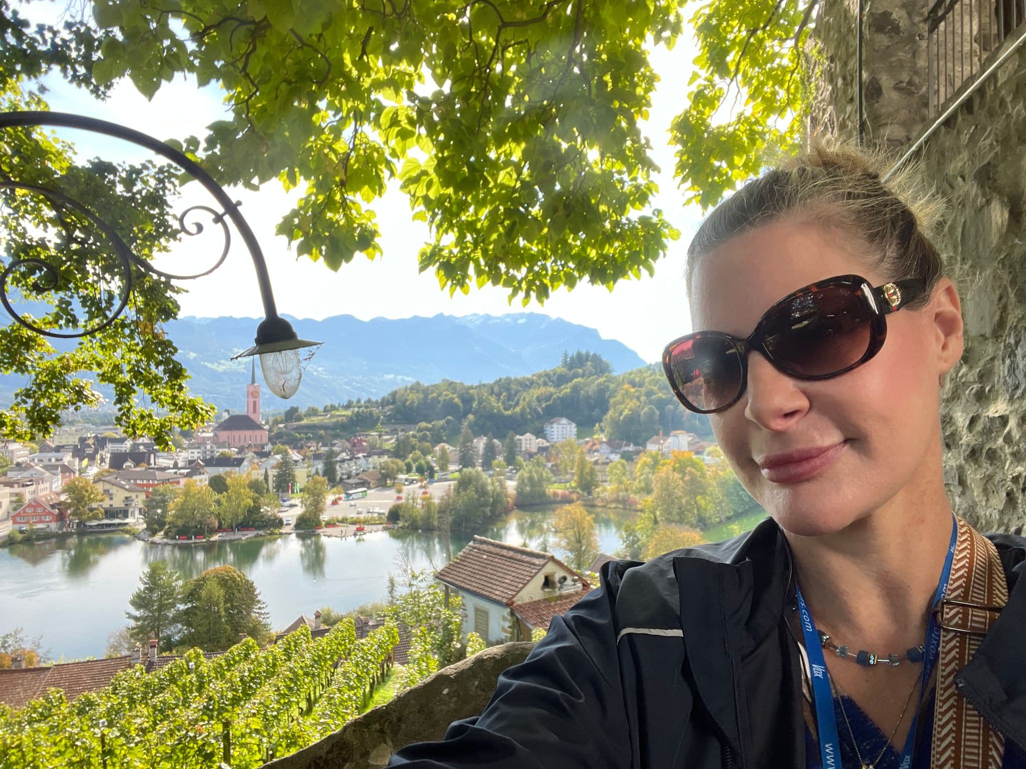 Riverside town and lake of Grabs with mountains beyond, framed by tree leaves and a traveler taking a selfie, Grabs, Switzerland.