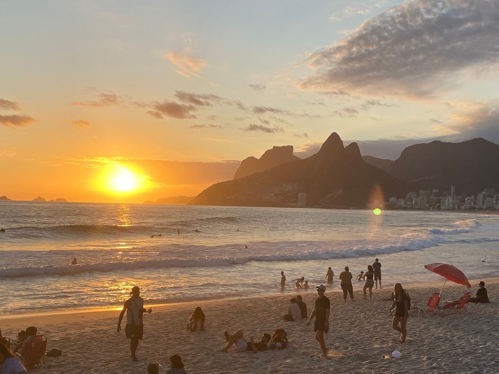Ipanema Beach at sunset with the Dois Irmãos mountains and people walking, sitting, and playing at the water's edge in Rio de Janeiro, Brazil.