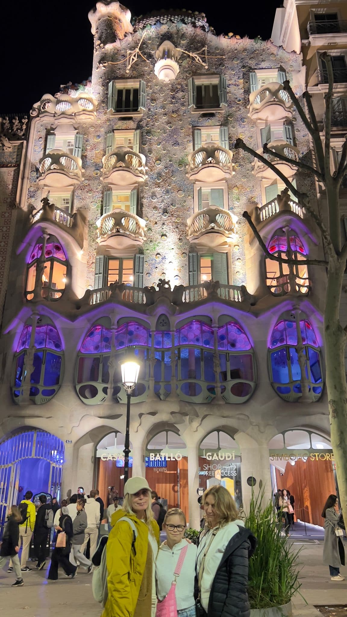 Casa Batlló lit at night with three travelers posing in front on a busy street, Barcelona, Spain.
