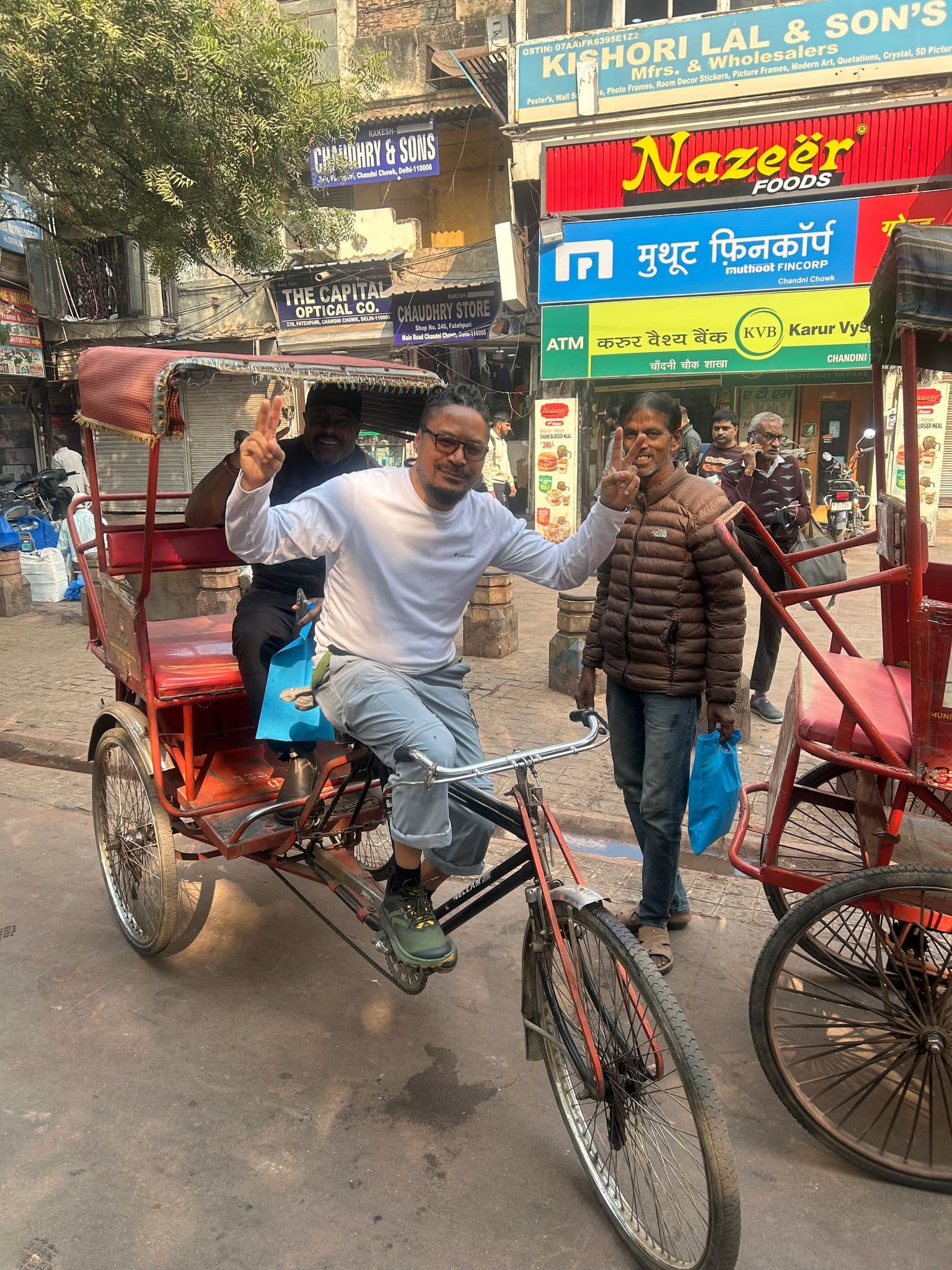 Man seated on a cycle rickshaw flashing peace signs in a busy Chandni Chowk street, Old Delhi, India