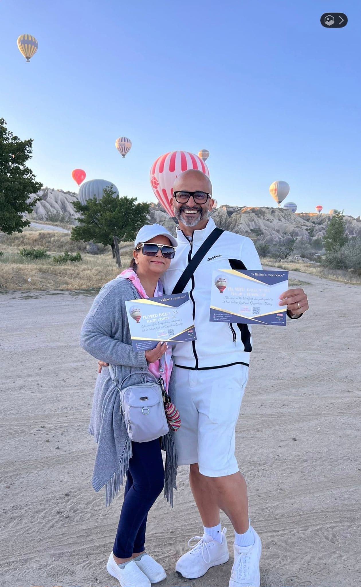Hot air balloons rising over the Fairy Chimneys in Cappadocia, Turkey, with a couple holding ride certificates in the foreground.