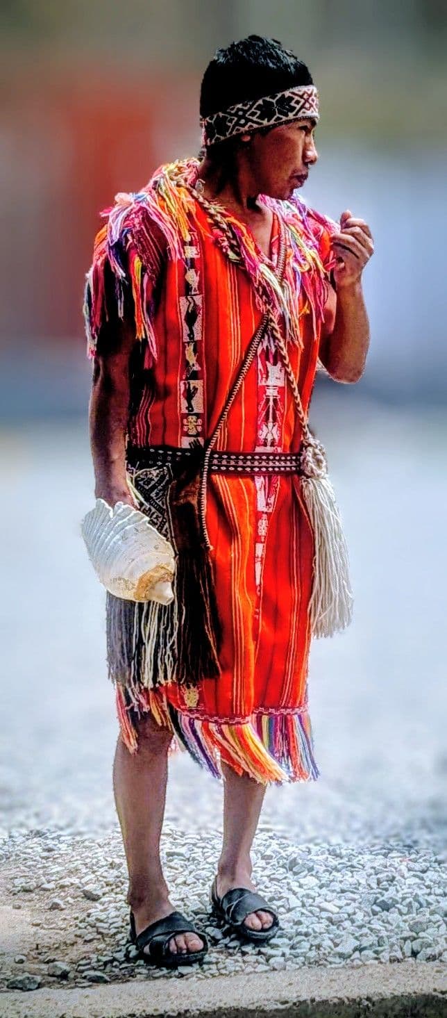 Man in red traditional Andean clothing standing on gravel near Machu Picchu, Peru, holding a shell-like object.