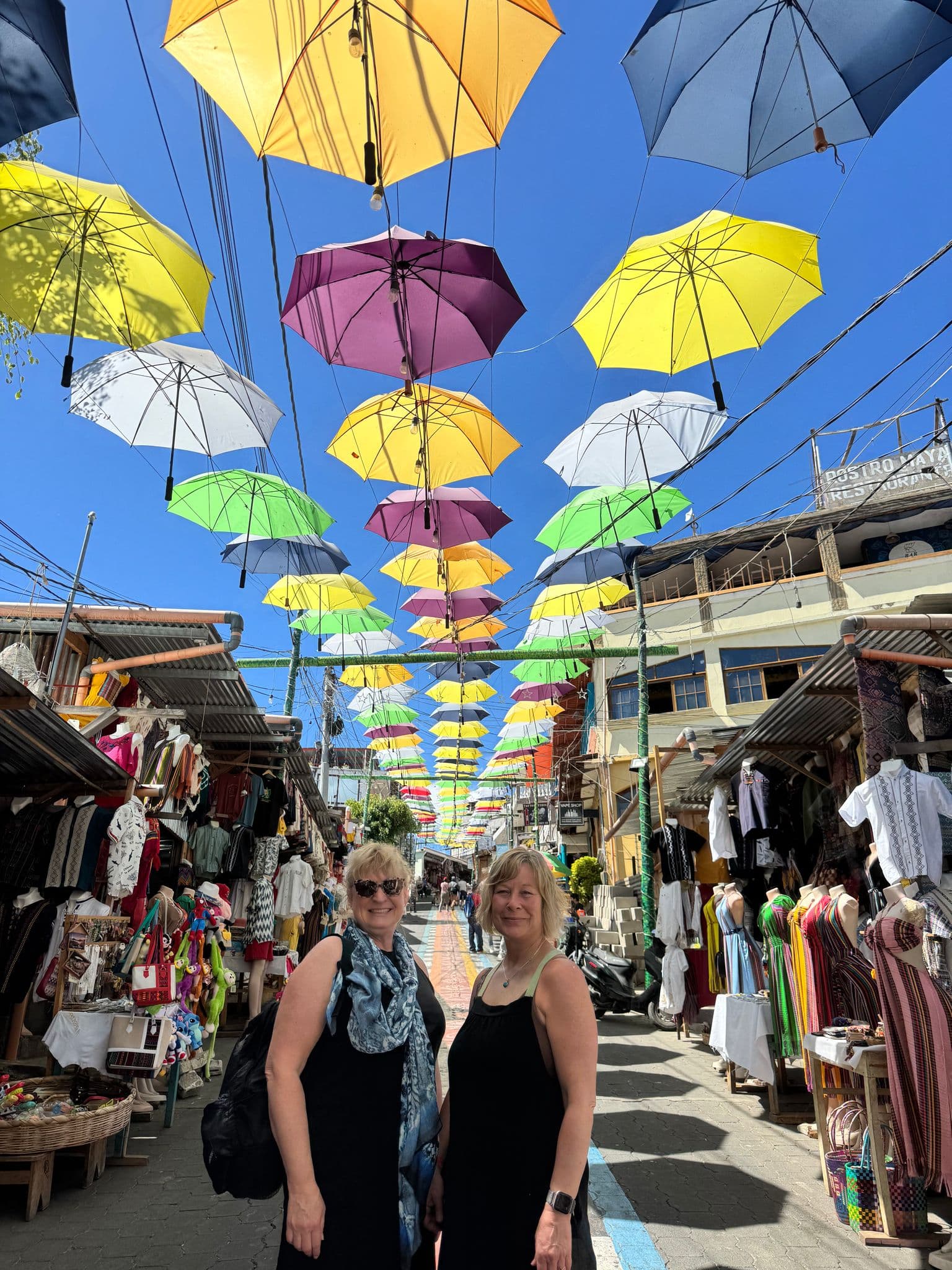 Colorful umbrellas suspended above a lively market street near Lake Atitlán, Guatemala, with two women posing in the foreground.