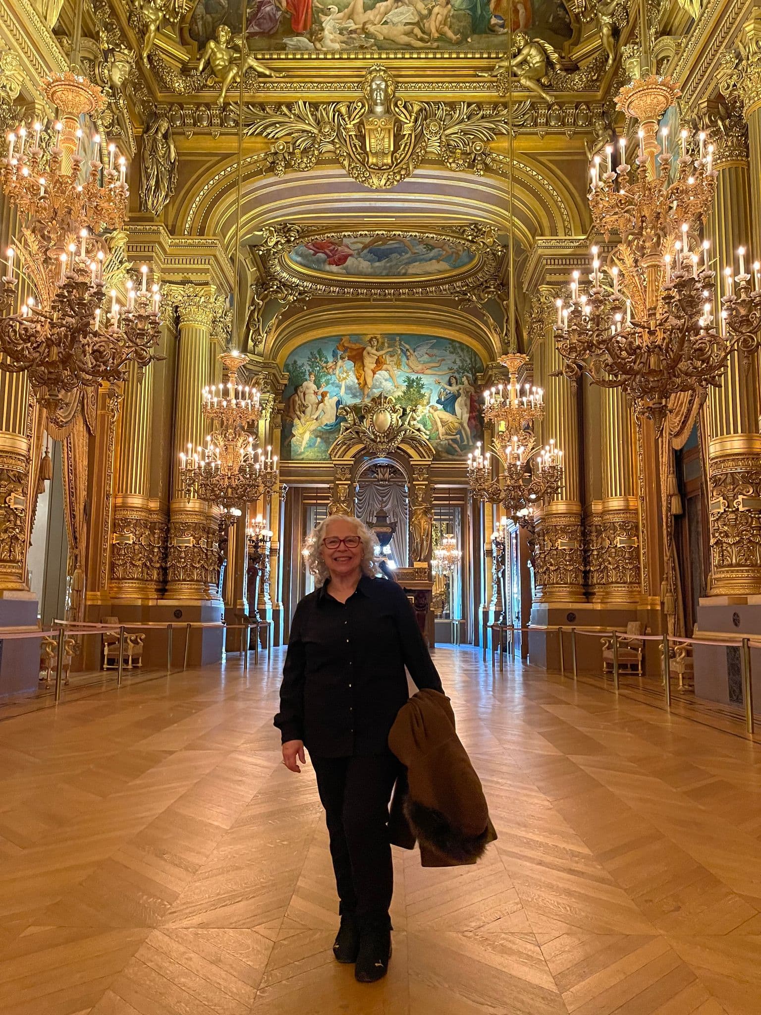 Palais Garnier interior with gilded chandeliers and painted ceiling; a person walks toward the camera holding a coat, Paris, France.