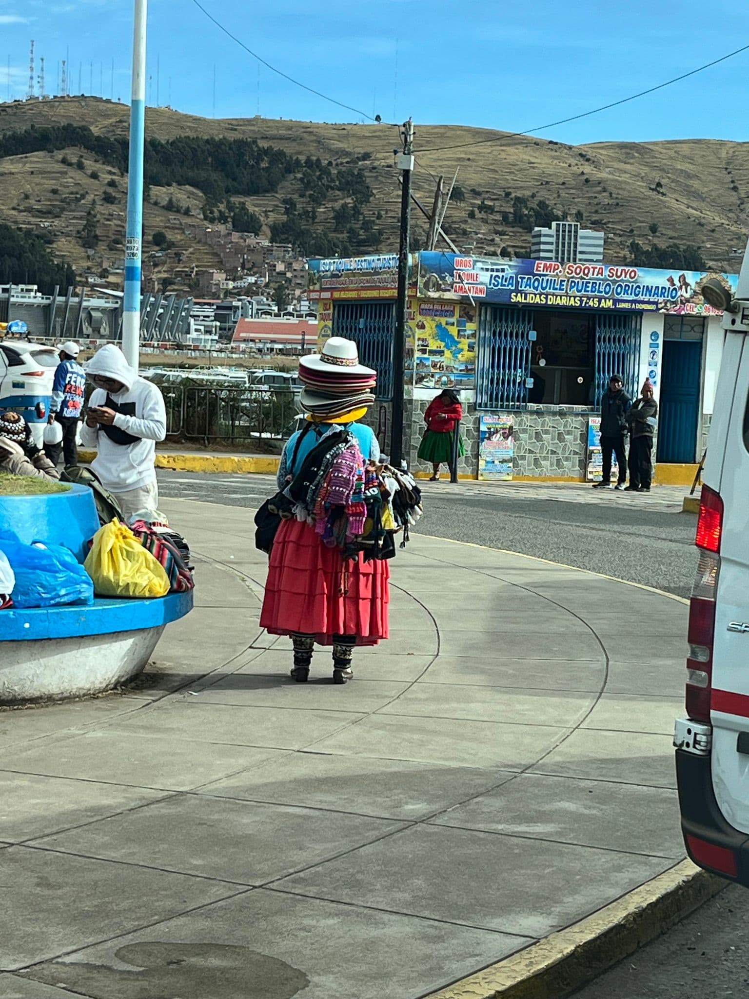 Street vendor wearing stacked traditional hats and colorful garments walking near the Puno ferry terminal to Isla Taquile, Puno, Peru.