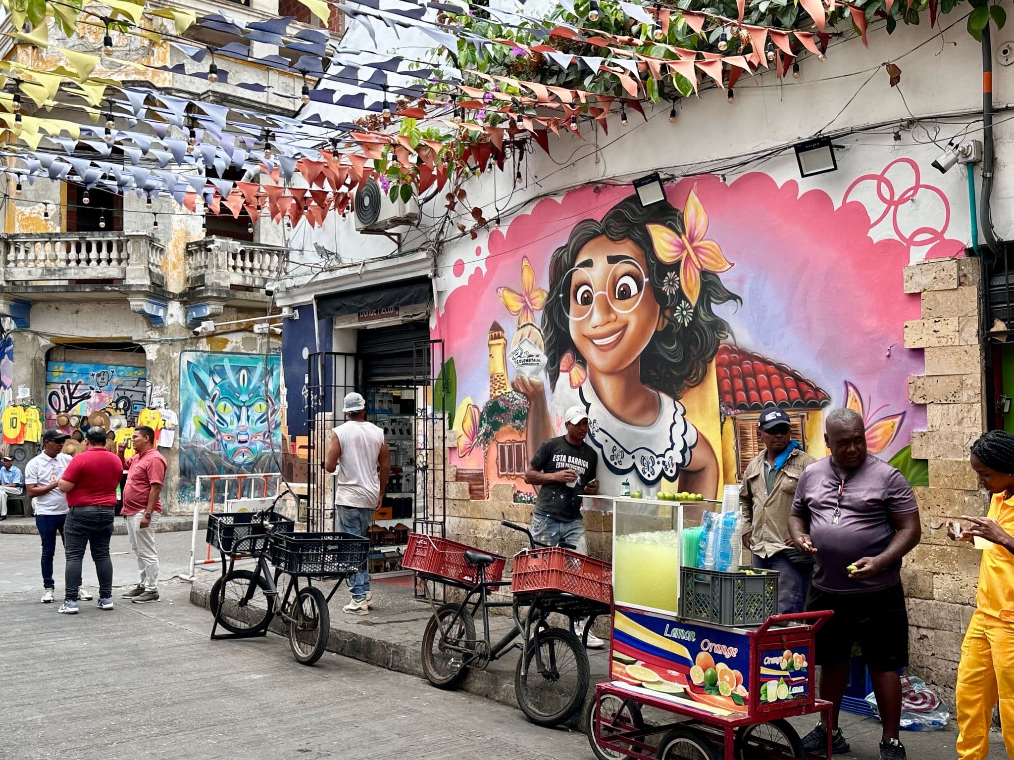 Large colorful mural of a smiling woman on a wall in Getsemaní, Cartagena, Colombia, with street vendors and pedestrians nearby.
