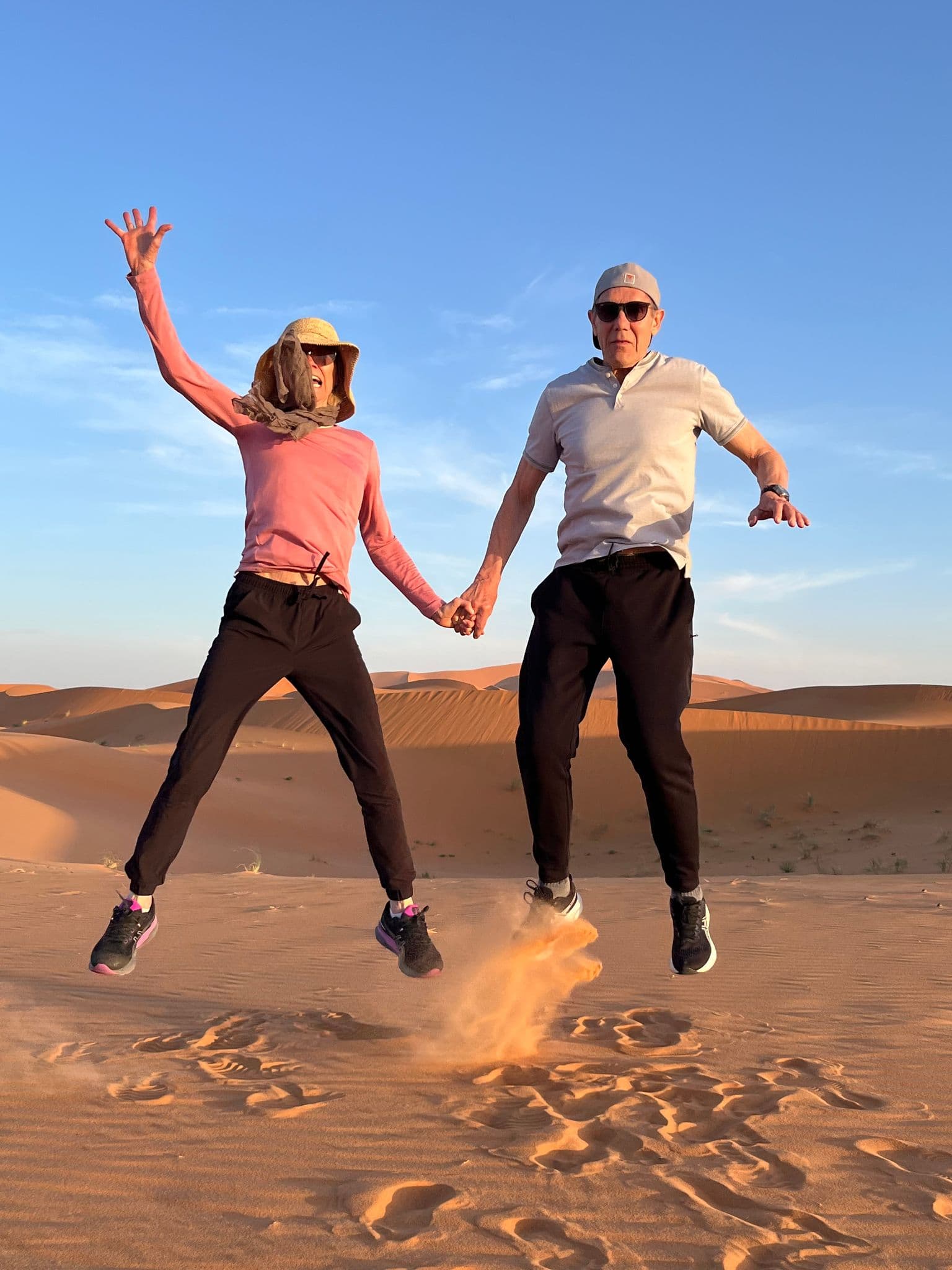 Two travelers holding hands jumping and kicking up sand on Sahara Desert dunes in Morocco.