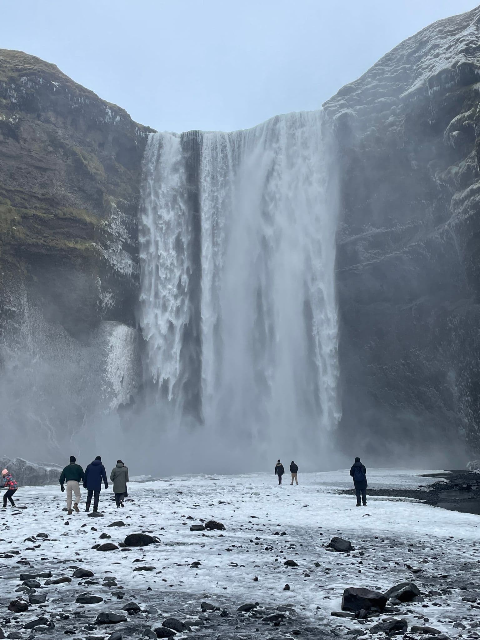 Skógafoss waterfall in Iceland cascading over a cliff while small groups of people walk on icy ground in front.