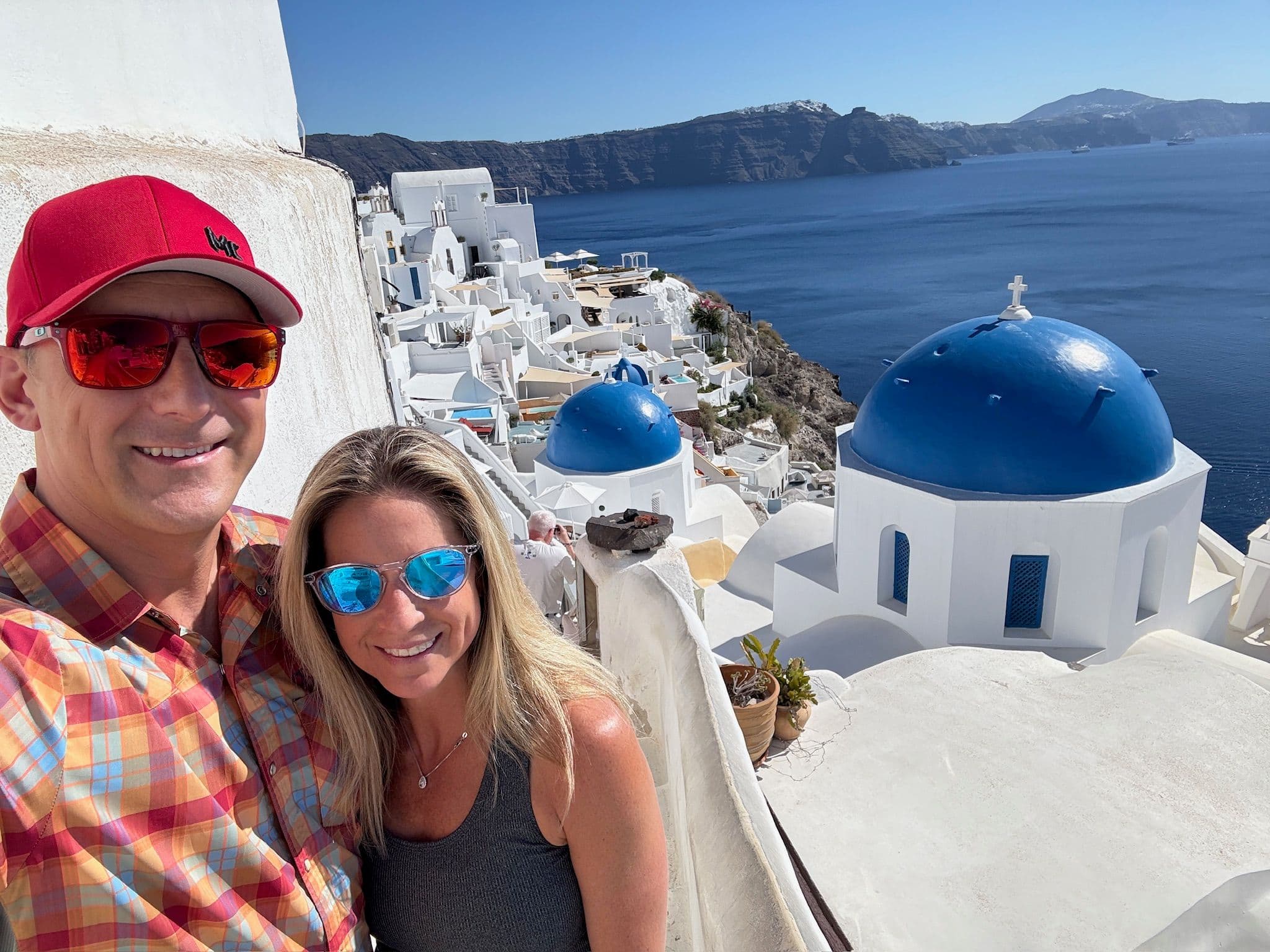 Blue-domed church in Oia, Santorini with a couple taking a selfie on a cliffside terrace overlooking the Aegean Sea, Greece.