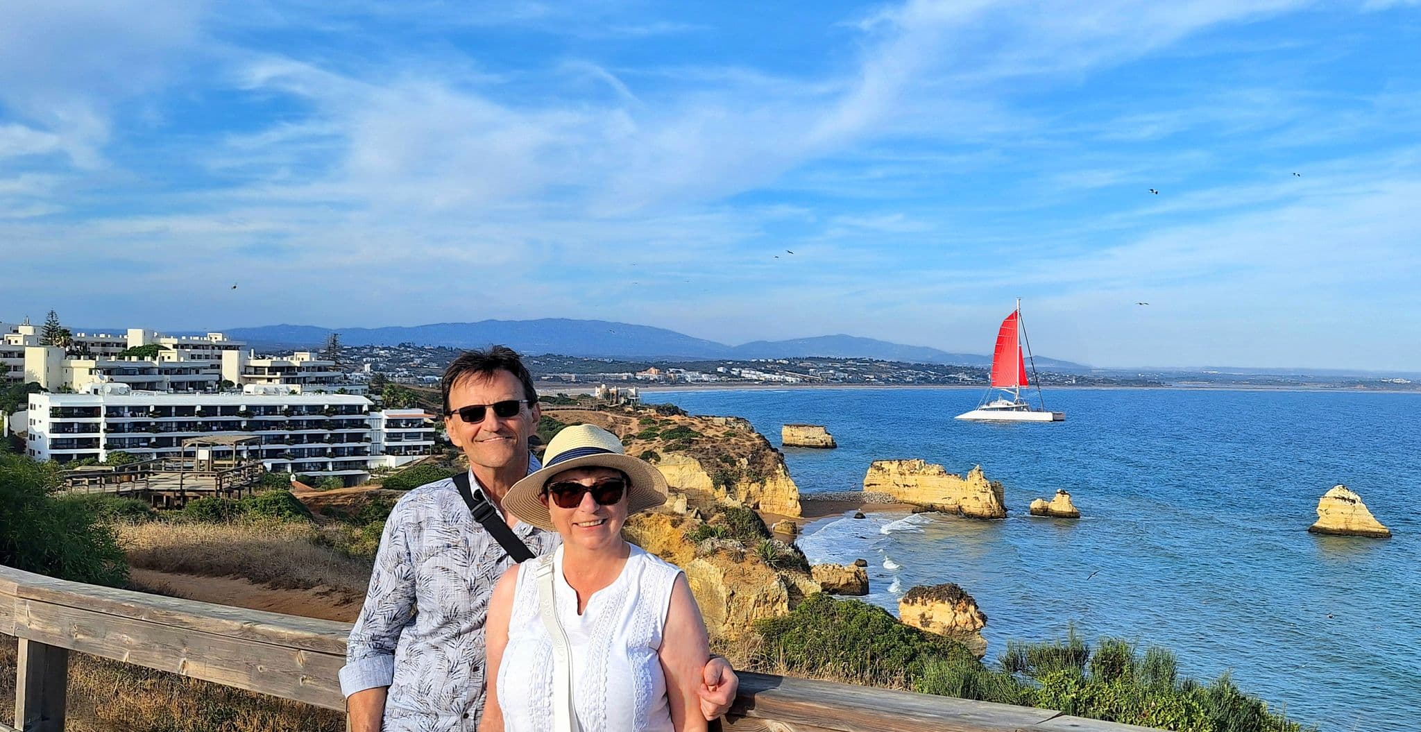 Ponta da Piedade cliffs and Atlantic with a red-sailed catamaran; a couple poses on a wooden viewpoint in Lagos, Portugal.