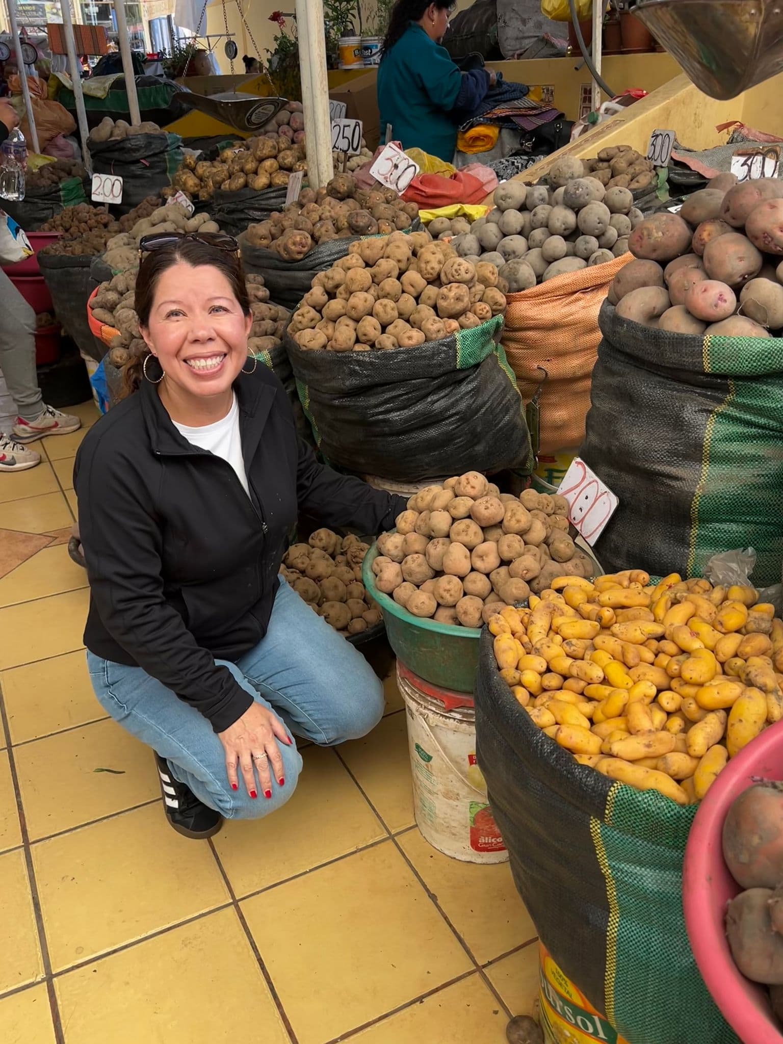 Piles of potatoes and a kneeling traveler at a local market in Cusco, Peru, browsing different varieties during a trip.