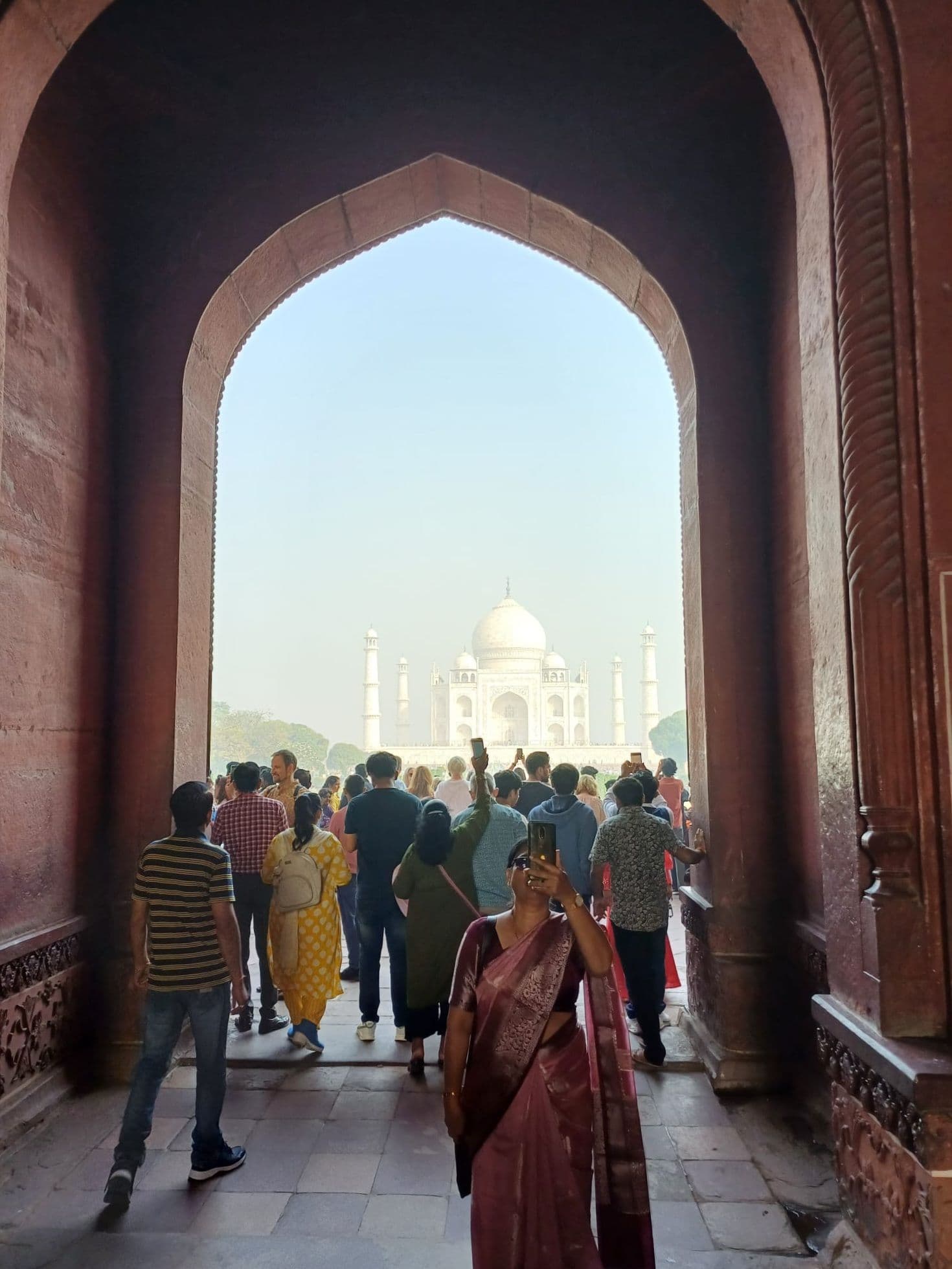 Taj Mahal framed by a red sandstone archway, crowds approaching it while a woman in a pink sari takes a selfie, Agra, India.