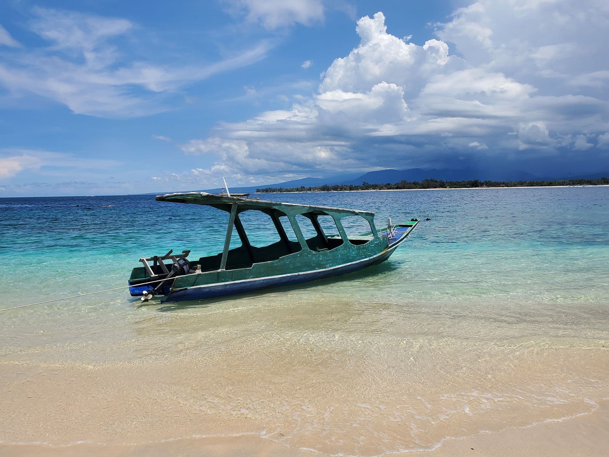 A green wooden boat beached in shallow turquoise water on Gili Trawangan, with Lombok island visible across the sea, Indonesia.