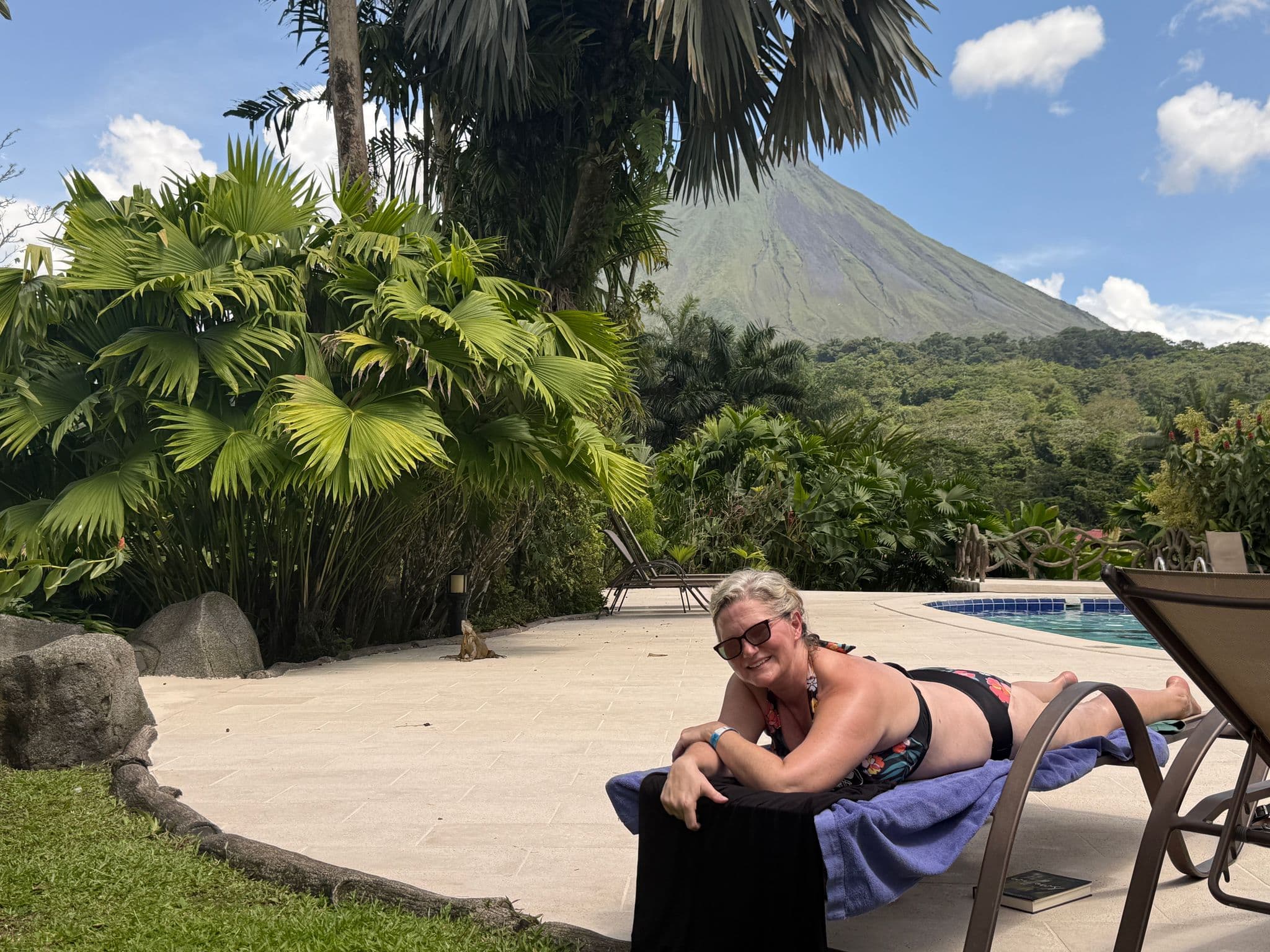 Arenal Volcano framed by tropical palms, with a woman sunbathing on a poolside lounge and an iguana on the patio, Costa Rica.