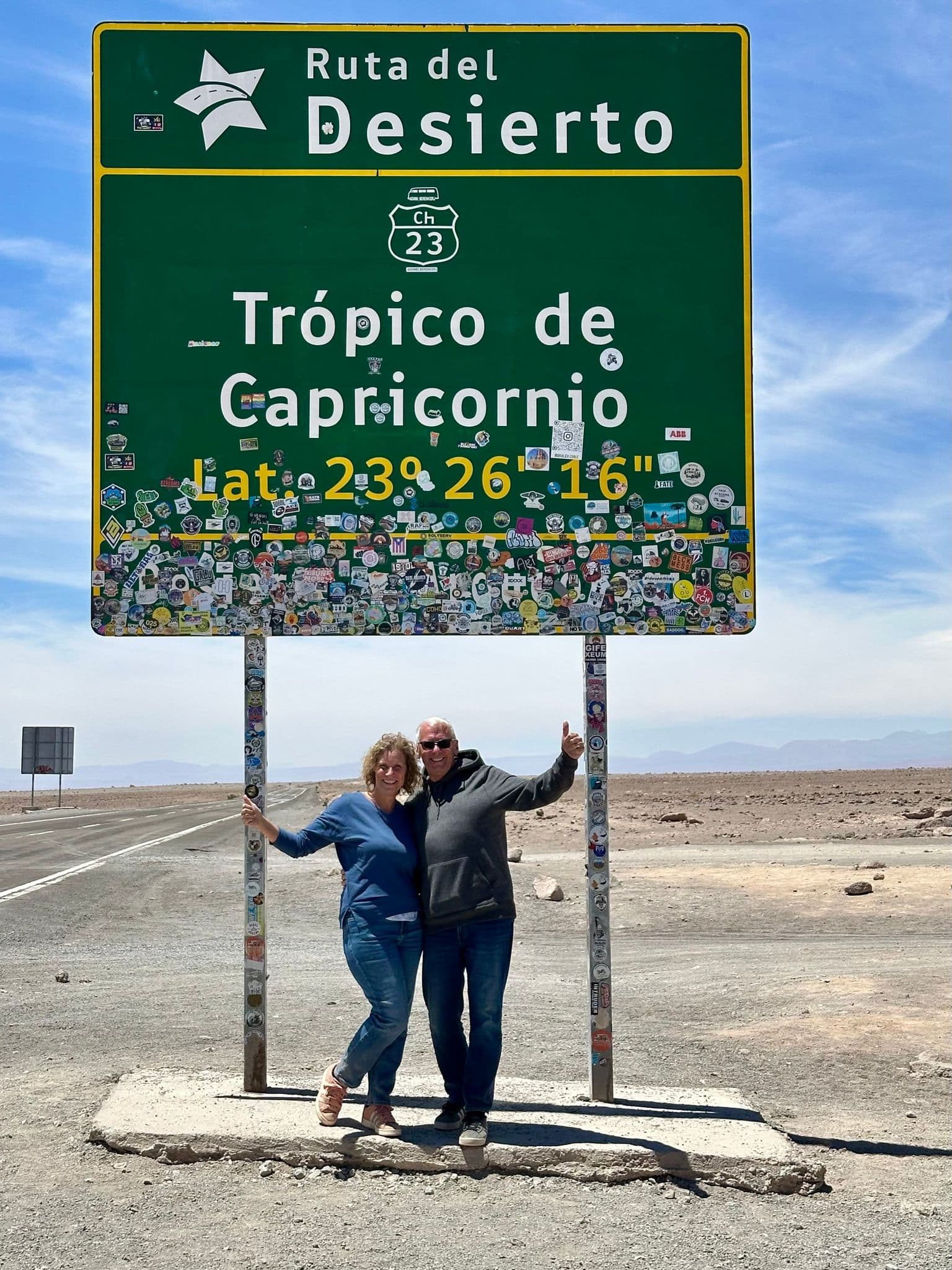Trópico de Capricornio road sign with two travelers posing beneath it in the Atacama Desert, Chile.
