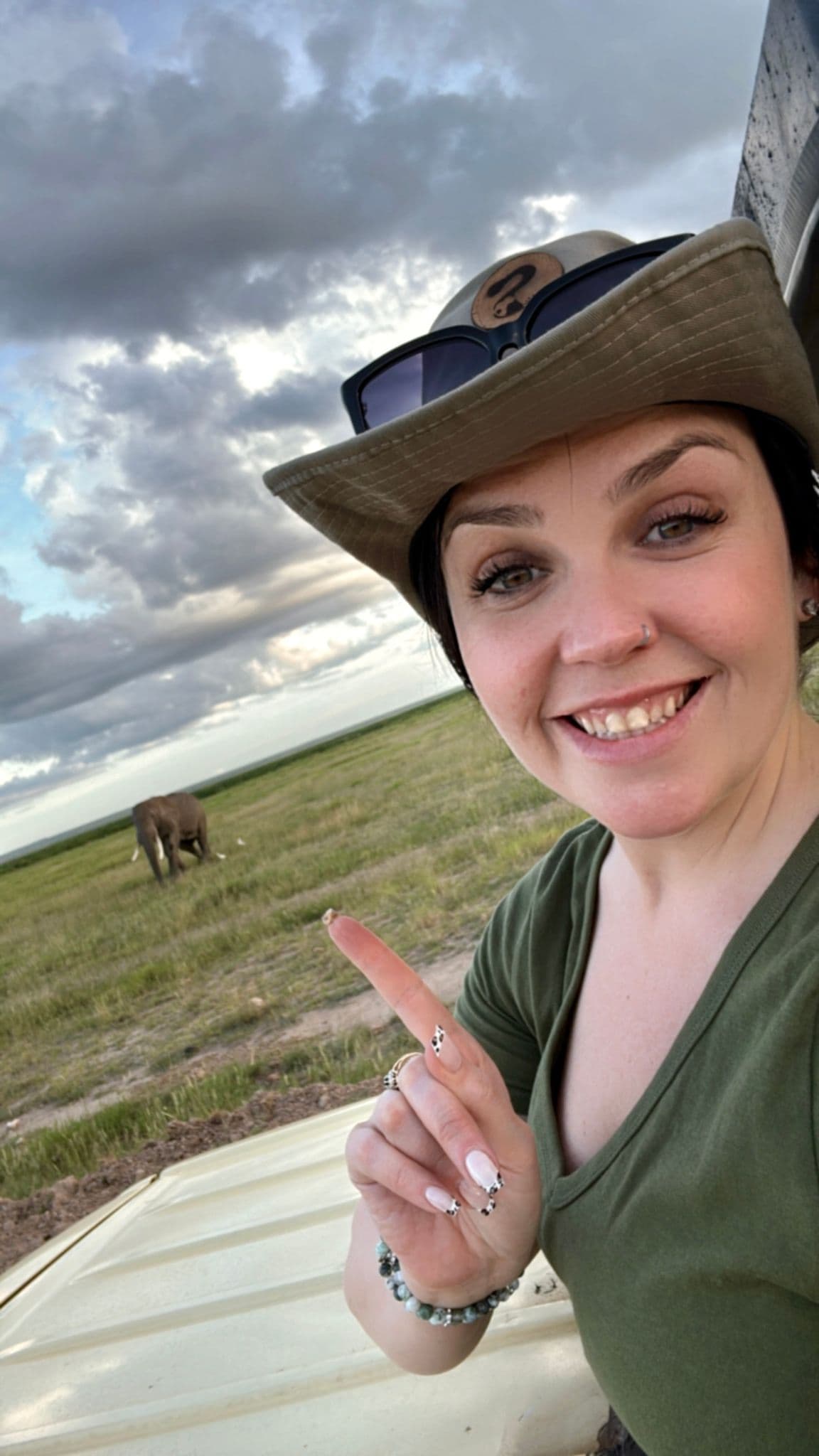Woman taking a selfie and pointing at an elephant grazing in open grassland during a safari trip.