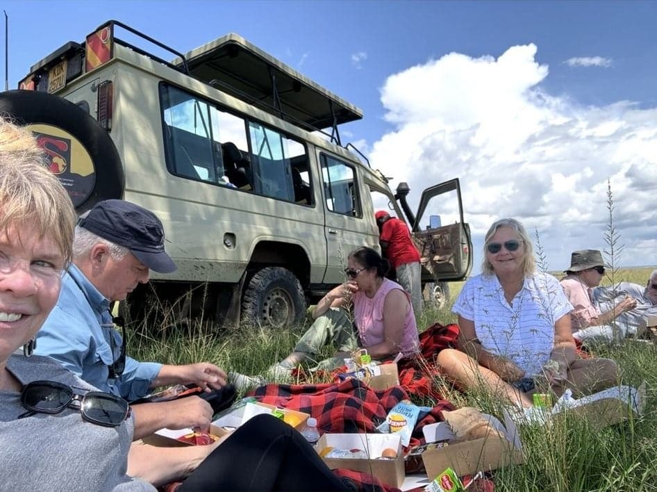 Safari vehicle and group picnic on grassland in Maasai Mara, Kenya, travelers sitting on a blanket eating lunch under a blue sky.