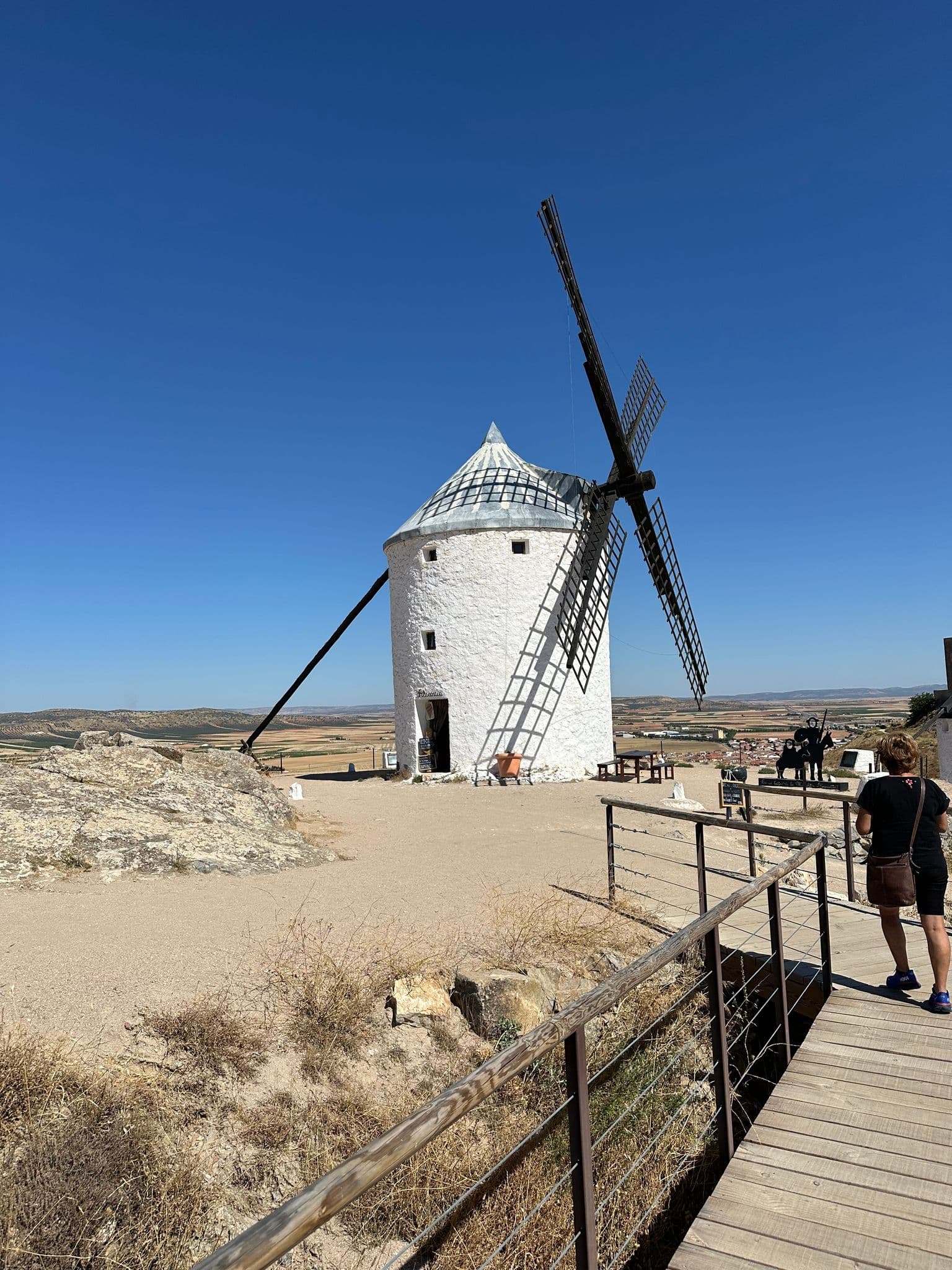 White La Mancha windmill with wooden sails under a clear blue sky, a tourist walking on a boardwalk, Castilla-La Mancha, Spain.