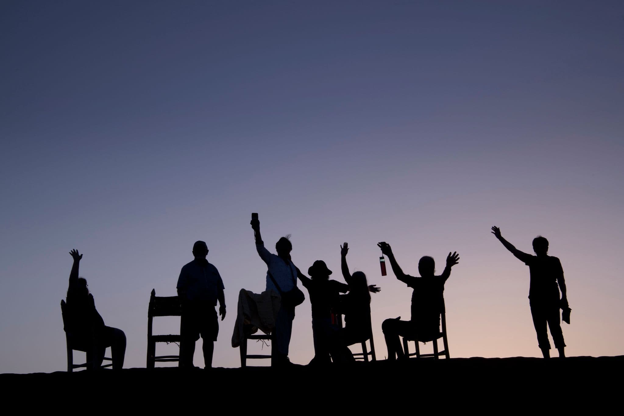 Sahara Desert sunset with silhouettes of travelers raising their hands and sitting on chairs on a dune during a trip.