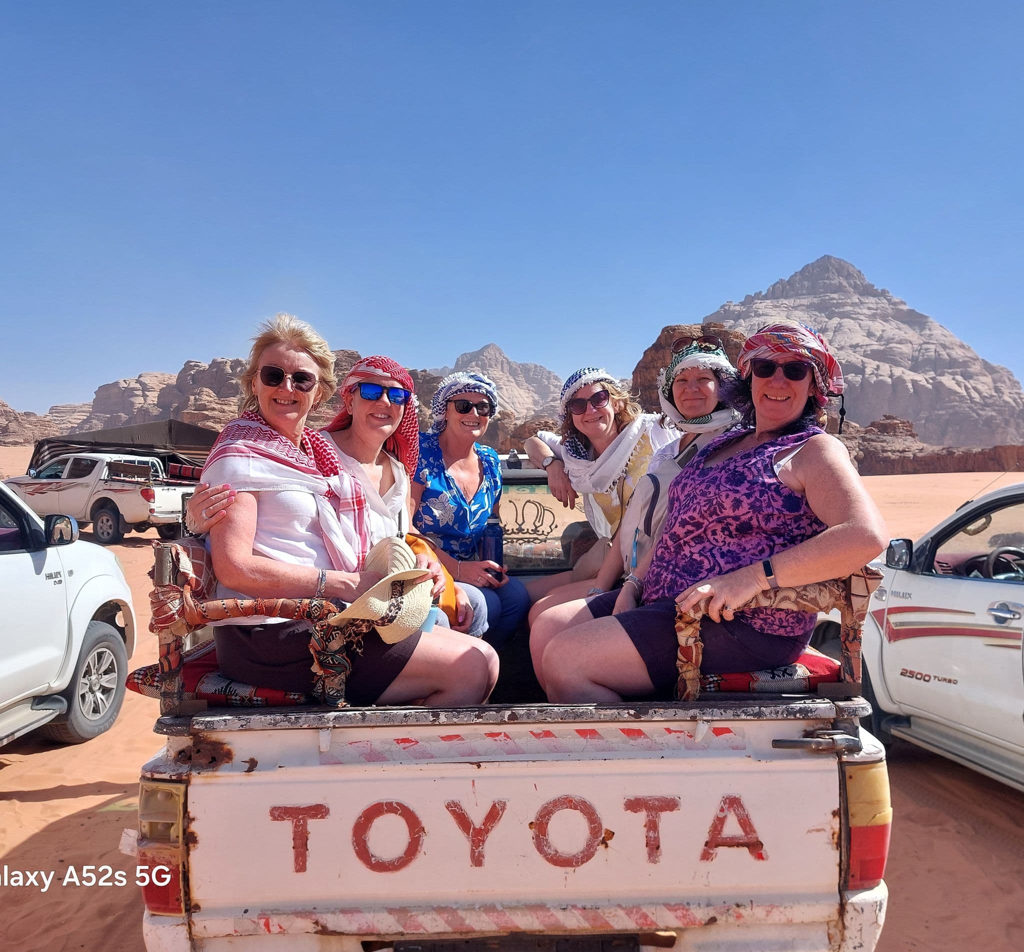Wadi Rum rock formation with six women in the back of a Toyota pickup wearing traditional scarves on a desert trip, Jordan.