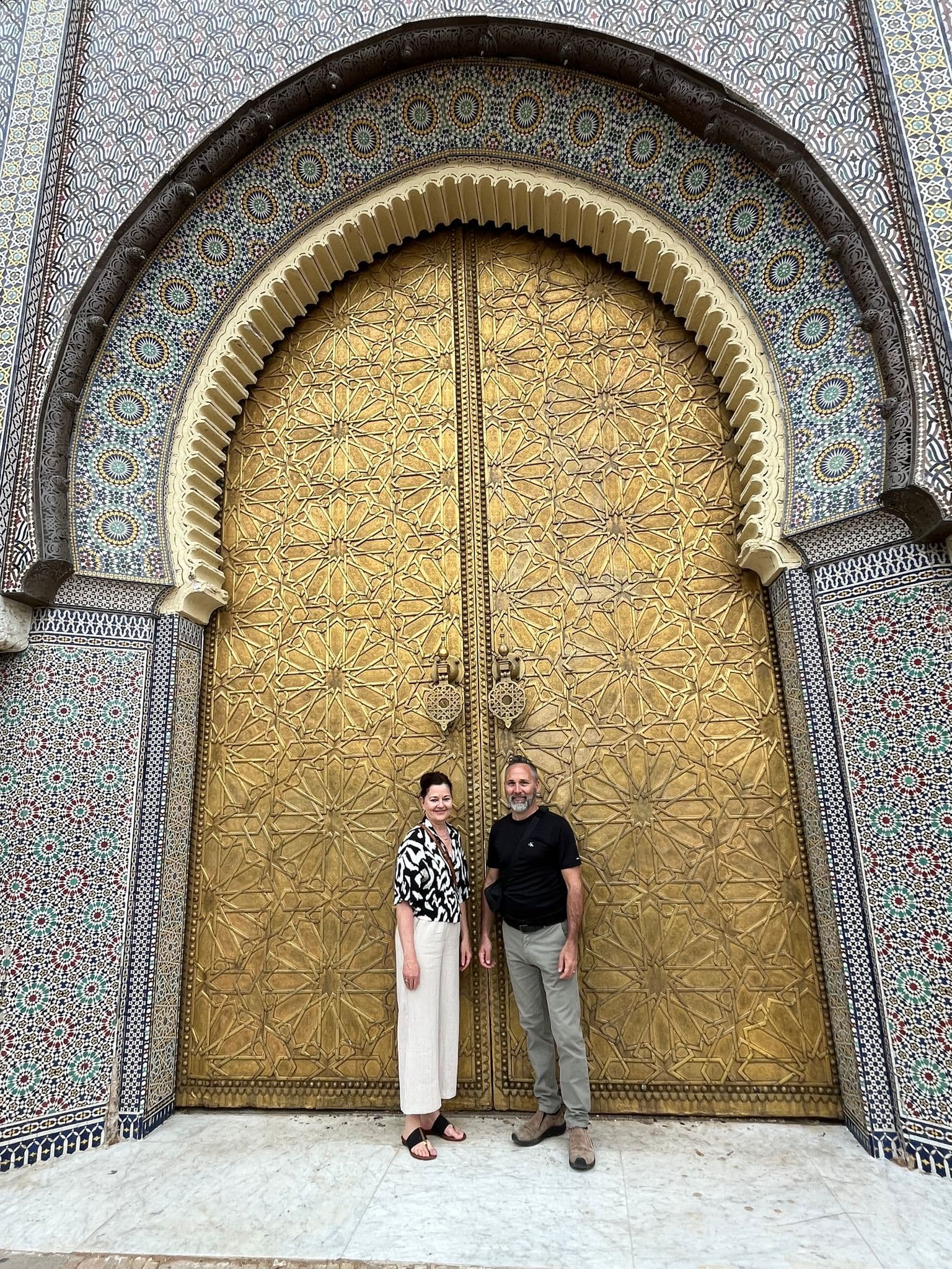 Golden doors of the Royal Palace (Dar el-Makhzen) in Fez, Morocco with two travelers standing beneath the tiled arch