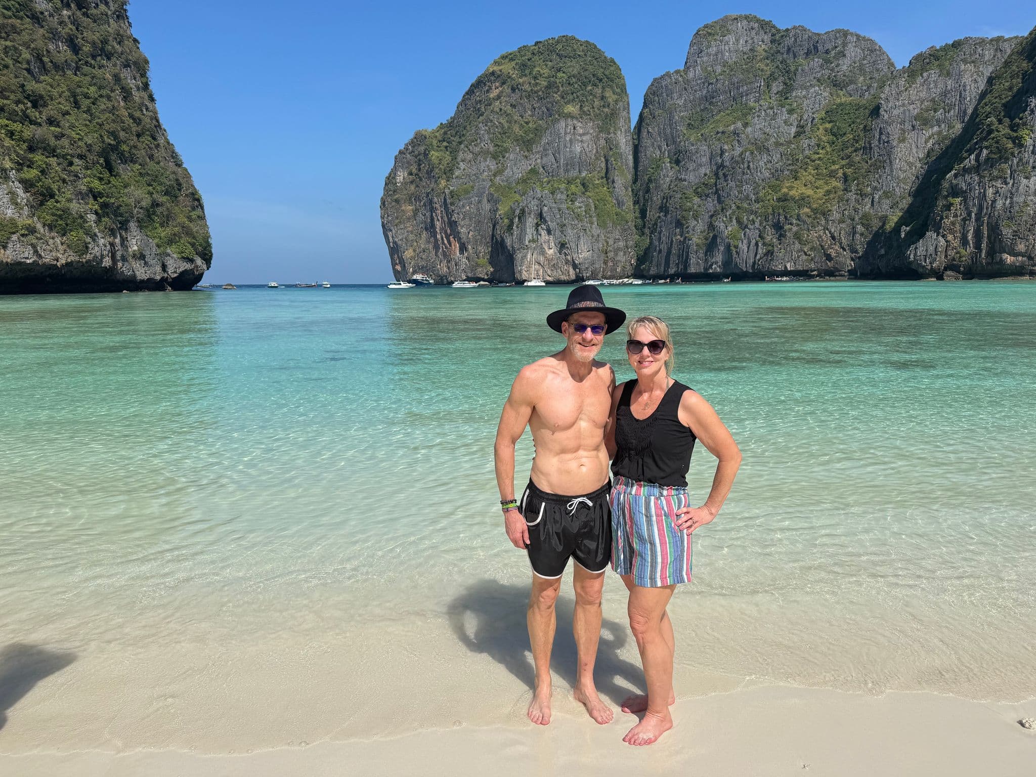 Maya Bay limestone cliffs and turquoise water with a couple standing on the sandy shore in Phi Phi Islands, Phuket, Thailand.