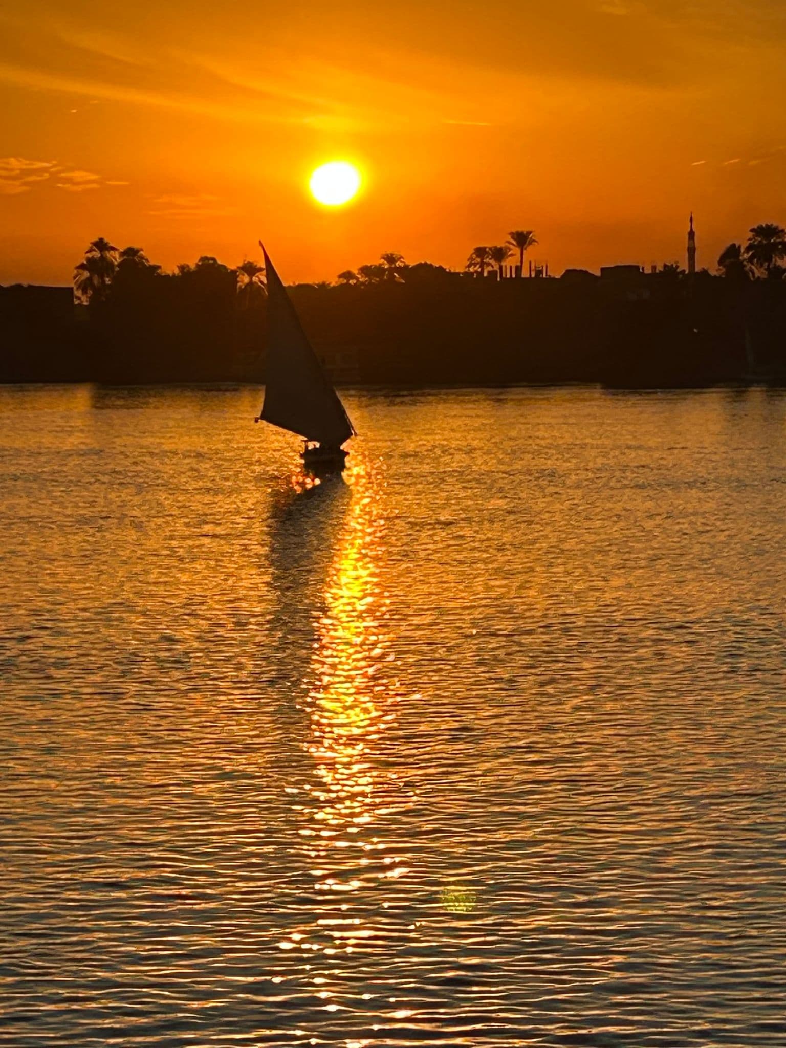 Nile River at sunset with a felucca sailing, the sun reflecting on the water and a silhouetted shoreline in Egypt.