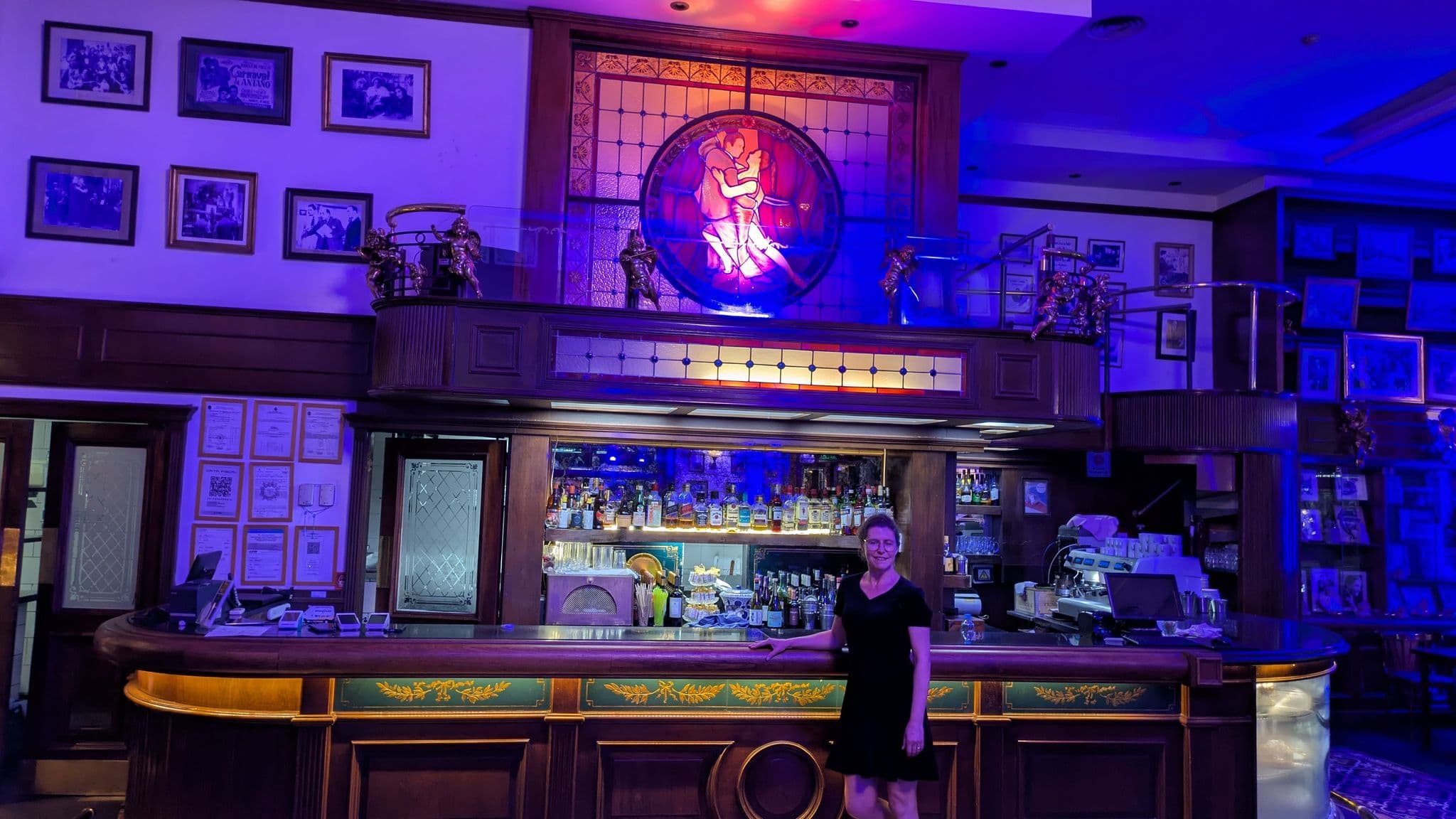 Stained-glass tango couple above a wood bar as a woman stands at the counter inside a historic tango cafe, Buenos Aires, Argentina.