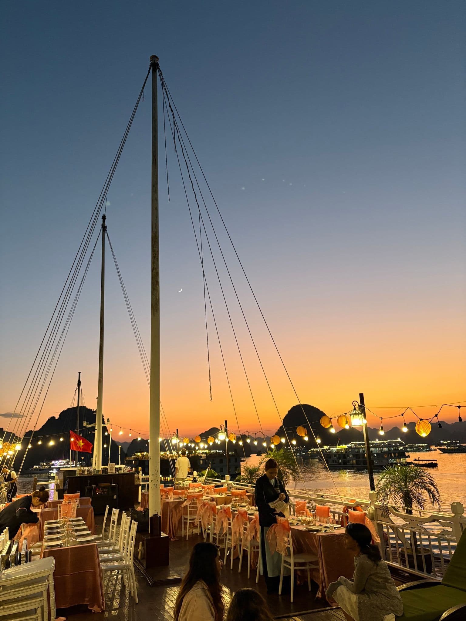 Ha Long Bay cruise deck with dining tables and string lights at sunset, karst islands and boats in Vietnam.