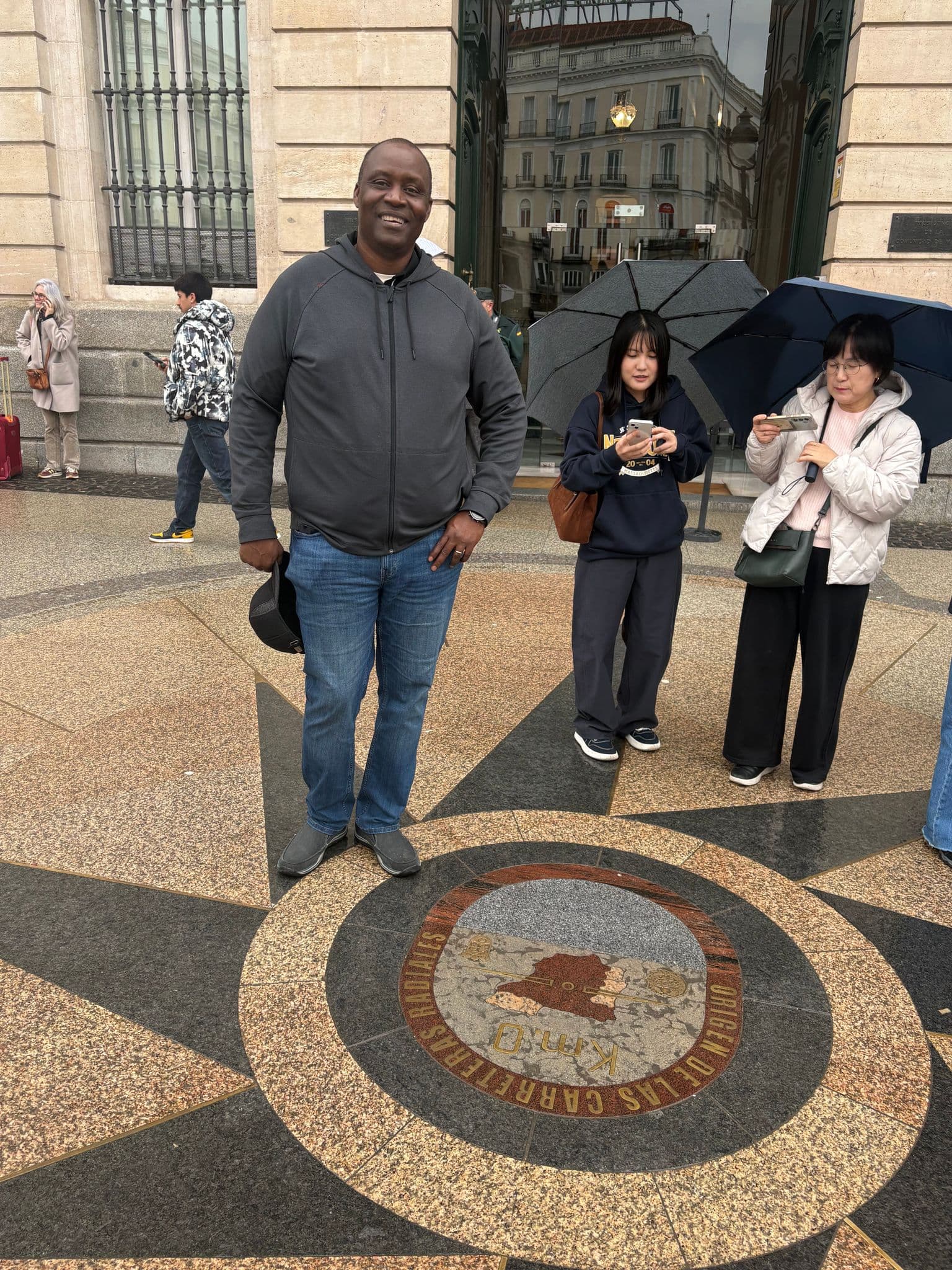 Kilómetro Cero (KM 0) pavement marker at Puerta del Sol with a smiling man standing nearby and tourists with umbrellas in Madrid, Spain.