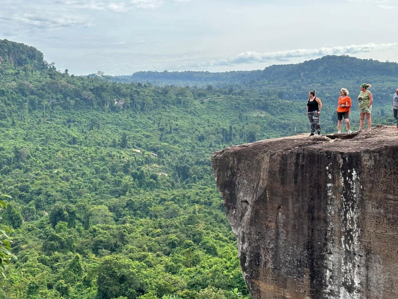 Rocky cliff edge with three people standing near the rim, overlooking a vast forested valley on a trip in Cambodia.