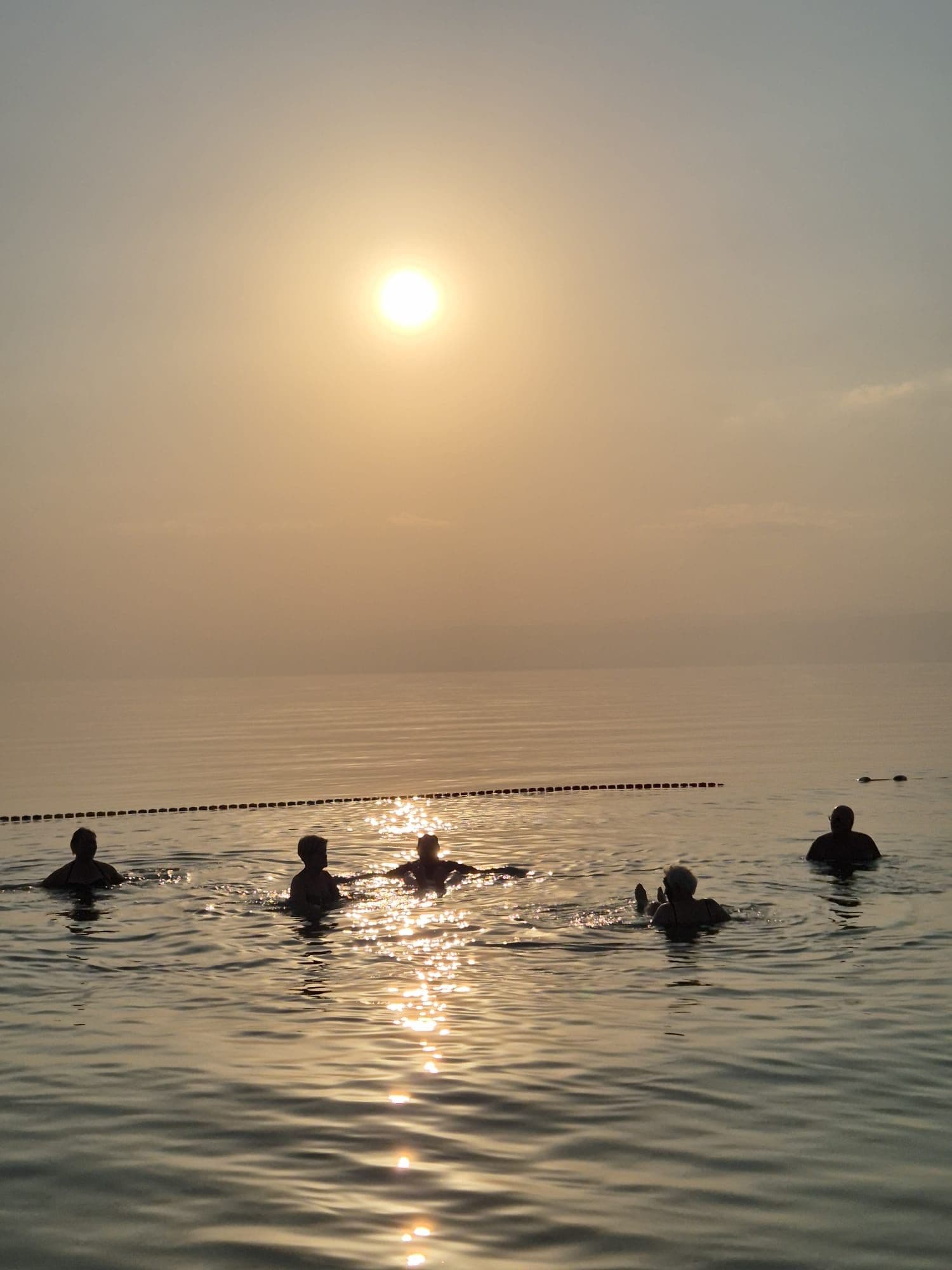 Sun over the Dead Sea in Jordan with five people floating and sunlight reflecting on the water.