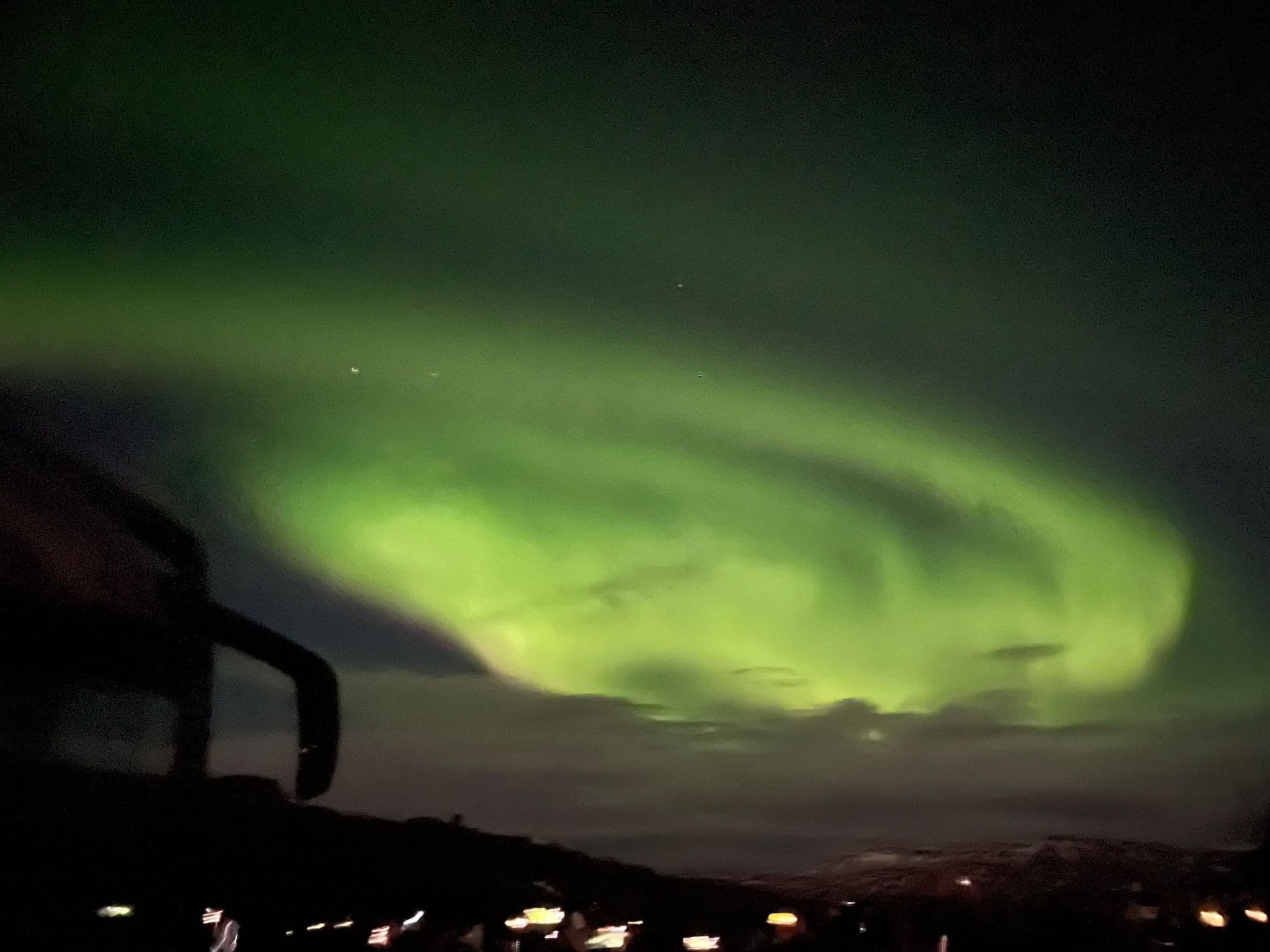 Aurora Borealis swirling green over a dark Icelandic landscape with distant town lights