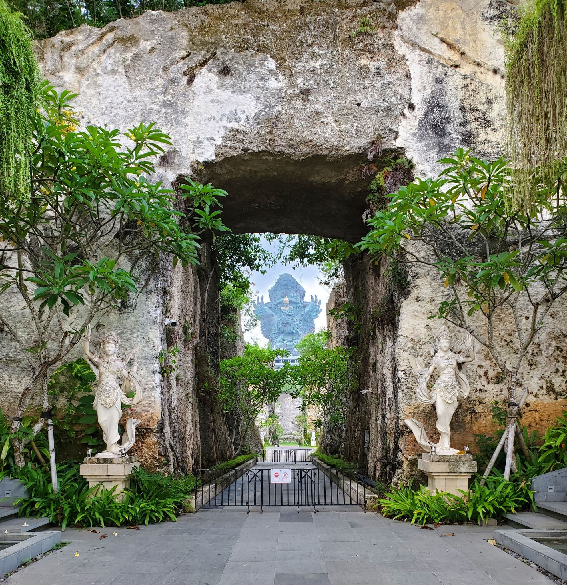 Garuda Wisnu Kencana statue framed by a natural rock archway with carved guardian statues at the park entrance, Bali, Indonesia