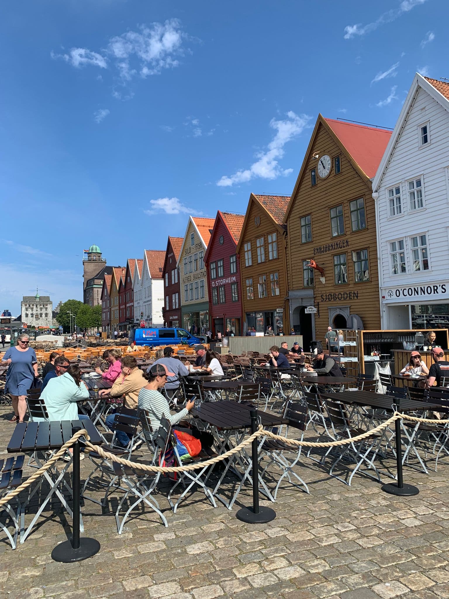 Row of colorful wooden Bryggen houses with outdoor café tables and people dining in Bergen, Norway.