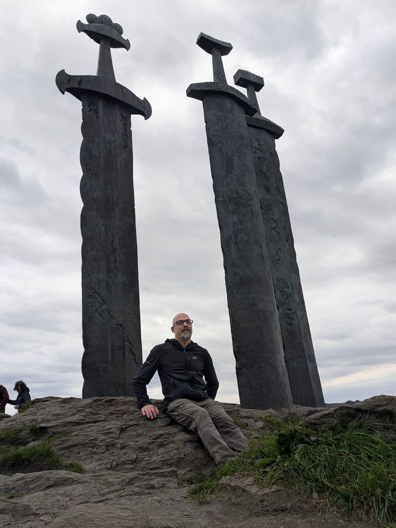 Sverd i fjell monument in Stavanger, Norway, with a man sitting on a rock before three large swords under a cloudy sky.