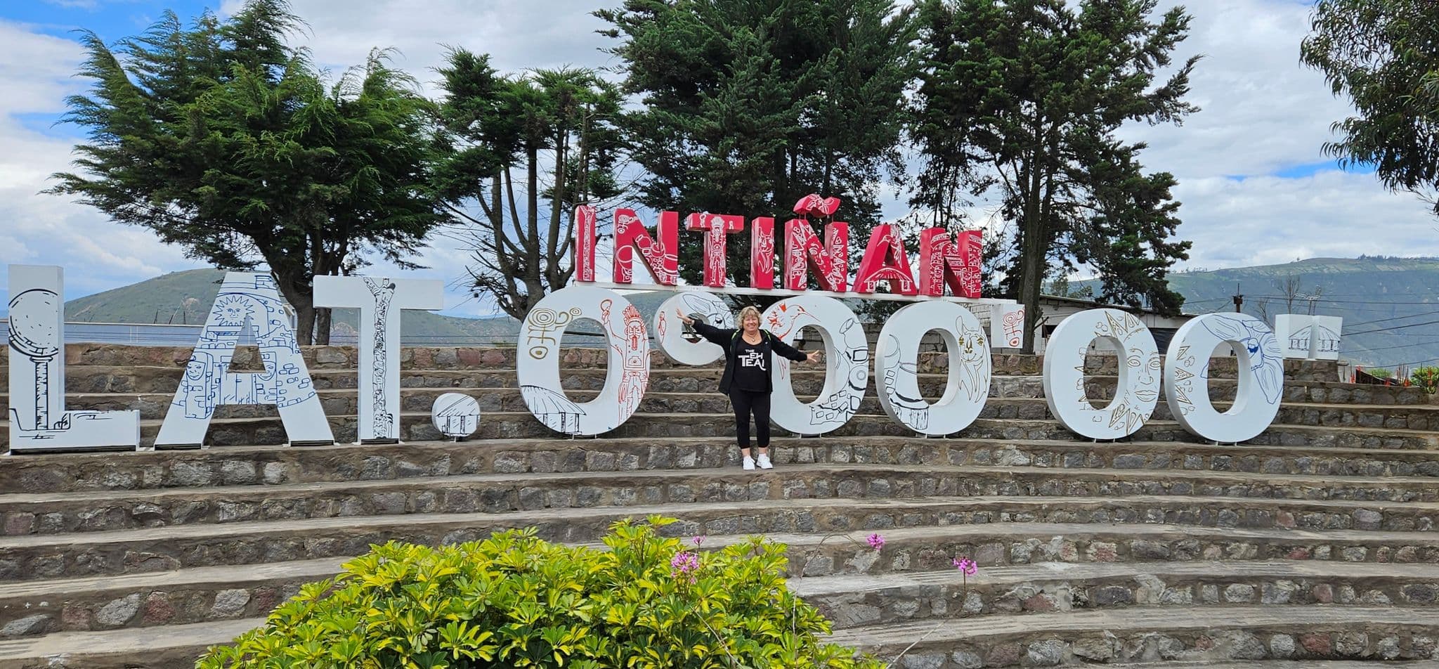 Large LAT 0°00'00 sign and red INTIÑAN letters with a traveler standing with arms outstretched at La Mitad del Mundo, near Quito, Ecuador.