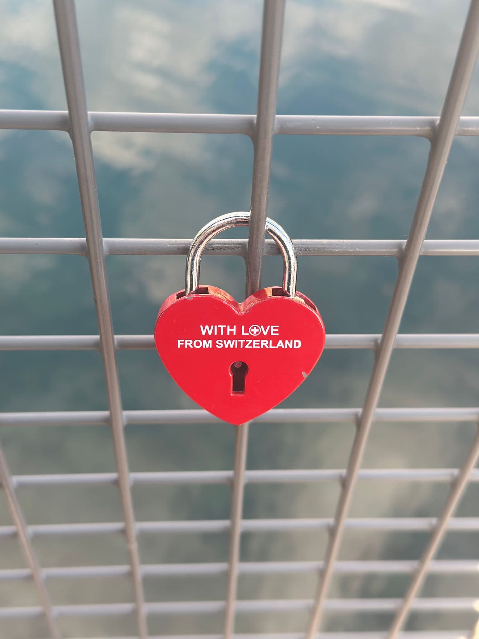 Red heart-shaped padlock reading "WITH LOVE FROM SWITZERLAND" locked to a metal bridge fence with water visible below, Switzerland.