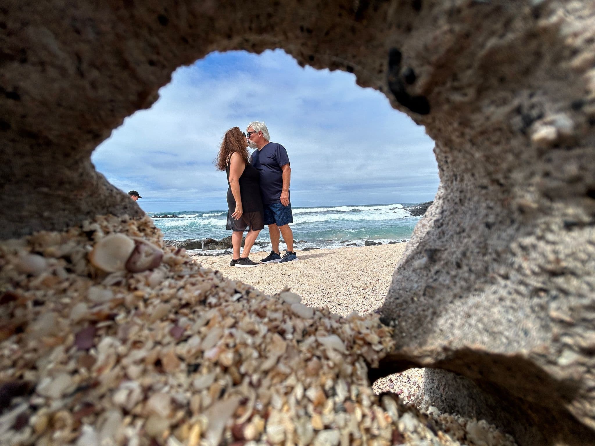Playa Los Frailes beach seen through a rock hole, a couple standing and kissing on the sand with waves behind them, Ecuador.
