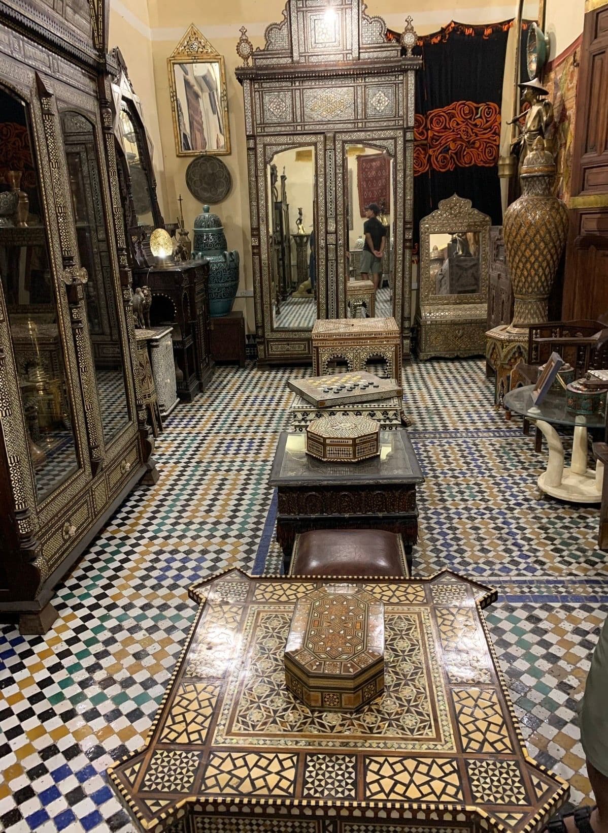 Inlaid wooden tables and colorful mosaic tiled floor inside an artisan shop in the Fez medina, Morocco, with a person in a mirror.