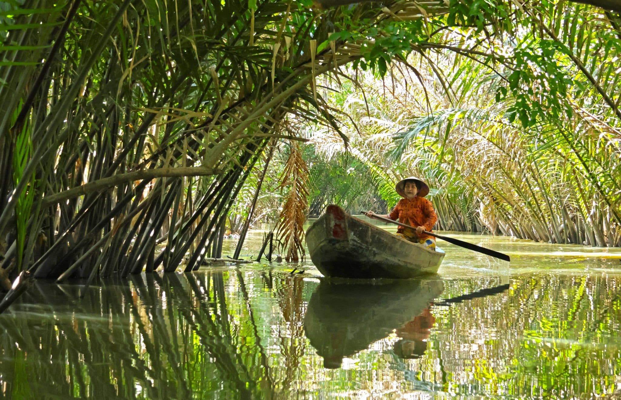 A small wooden boat with a person paddling through a palm-fringed channel in the Mekong Delta (Tre River), Vietnam.