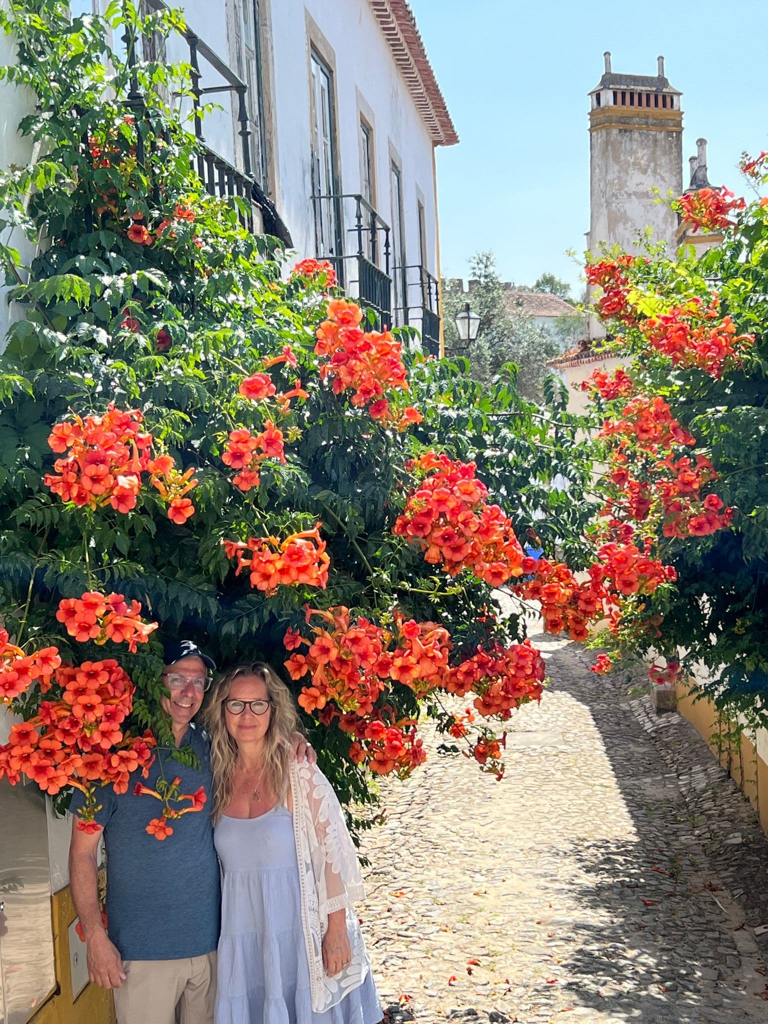 Óbidos street framed by orange trumpet-vine blooms, two travelers posing on a sunlit cobblestone lane, Portugal