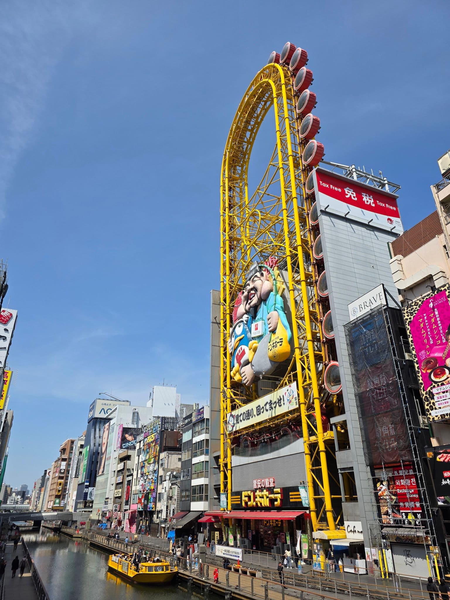 Ebisu Tower Ferris Wheel (Don Quijote) rising above Dotonbori canal with a yellow sightseeing boat and riverside walkers, Osaka, Japan.
