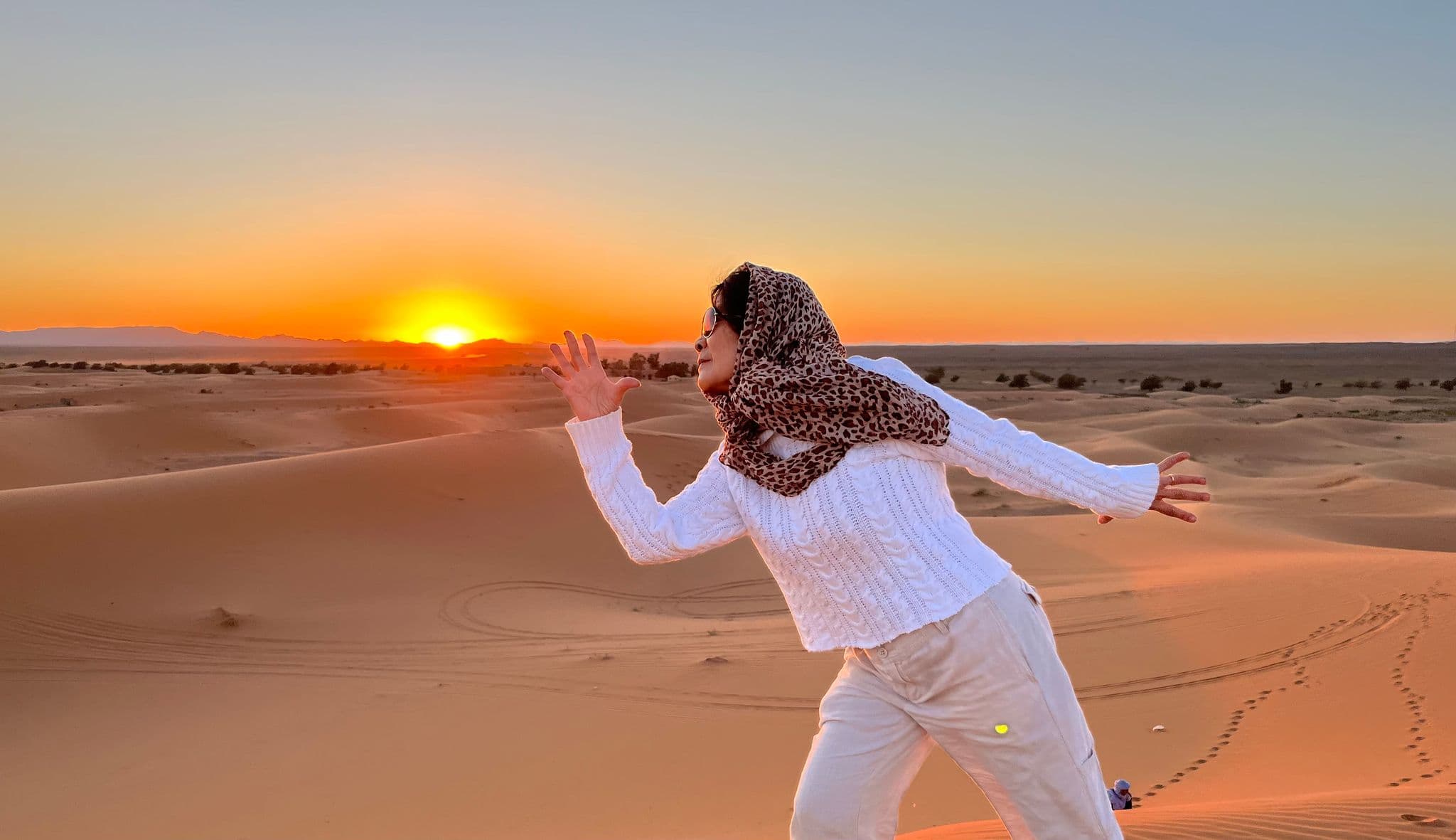 A woman in a patterned headscarf posing on sand dunes at sunset in the Sahara Desert.