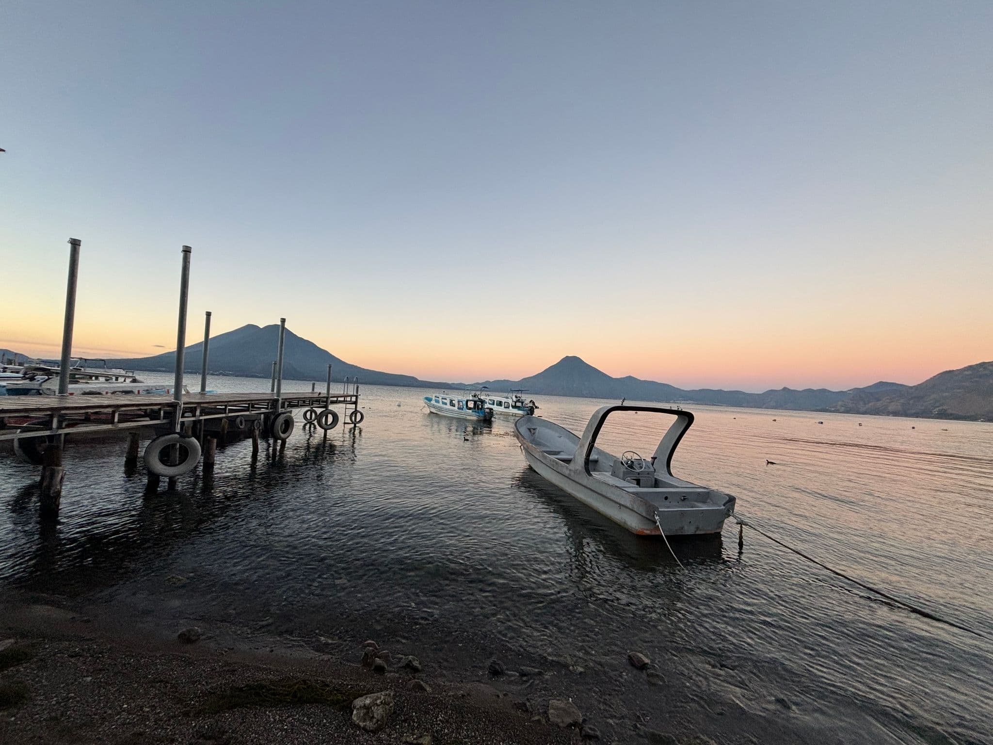 Lake Atitlán at sunset with a small motorboat tied to the shore and a wooden dock, volcanoes on the horizon, Guatemala.
